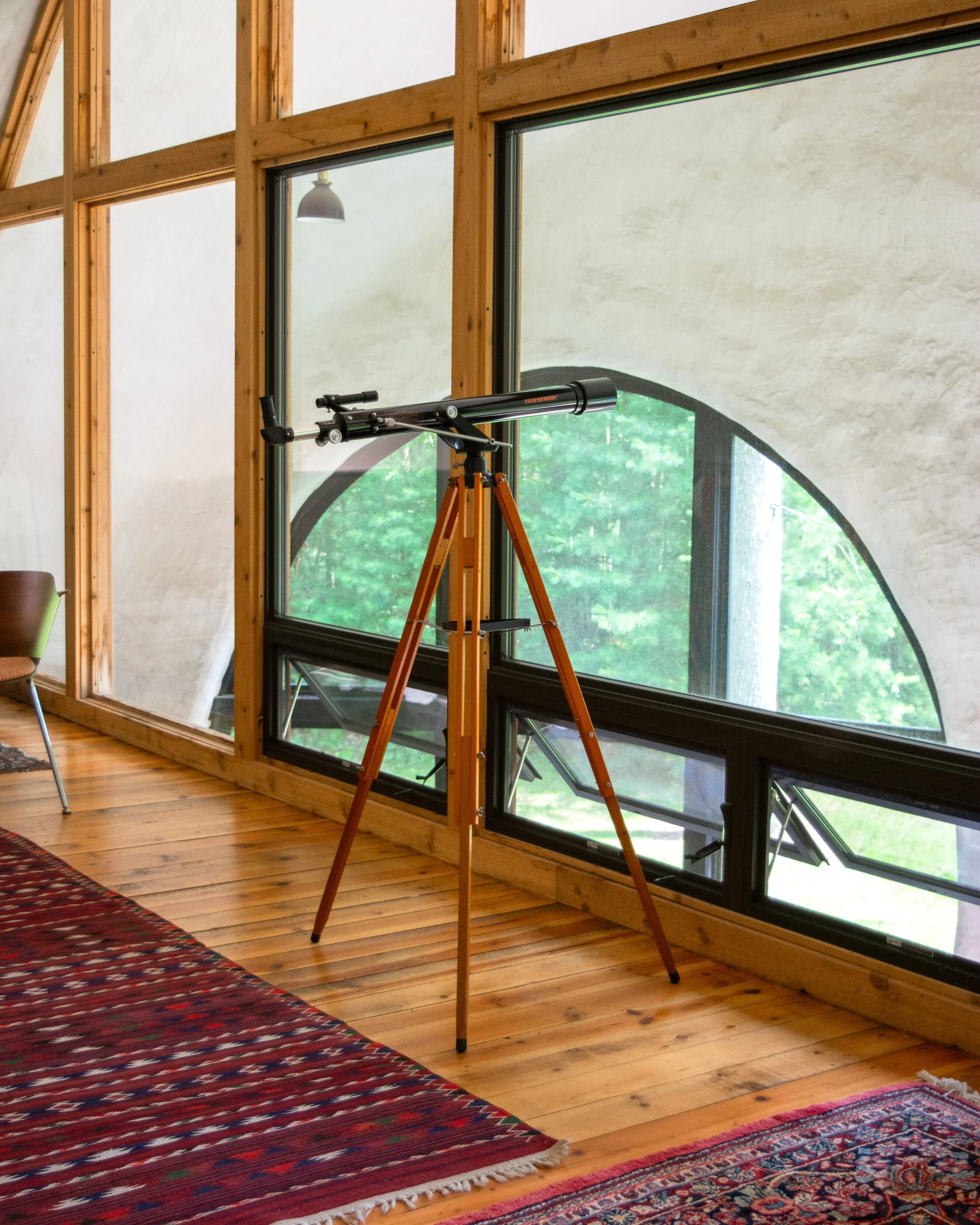 A viewing telescope on a wooden tripod inside a room with large windows and wooden floors.
