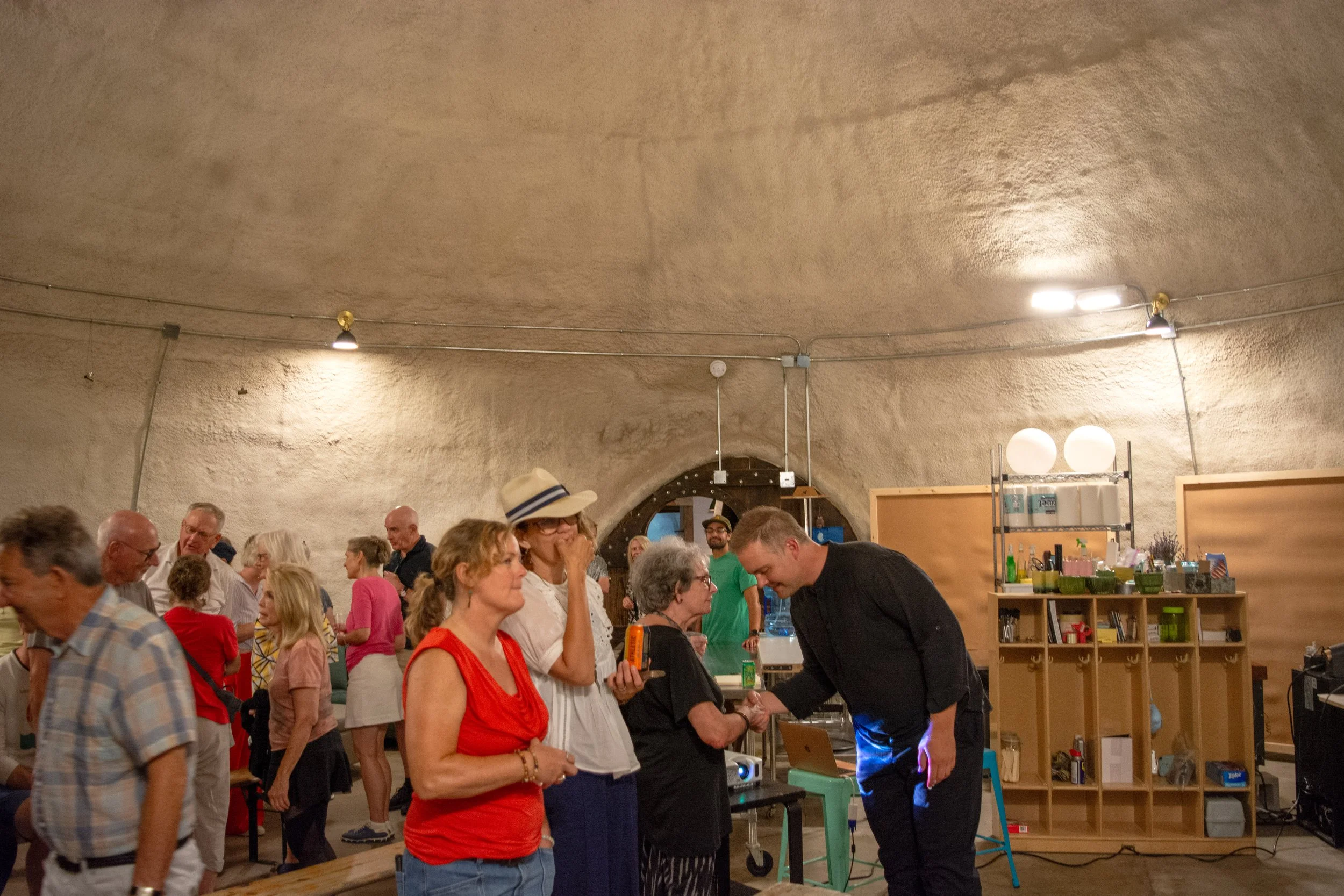 Group of people socializing at indoor event inside a large cave-like space with arched ceiling, some are standing and talking, others in line, with shelves and decorations in the background.