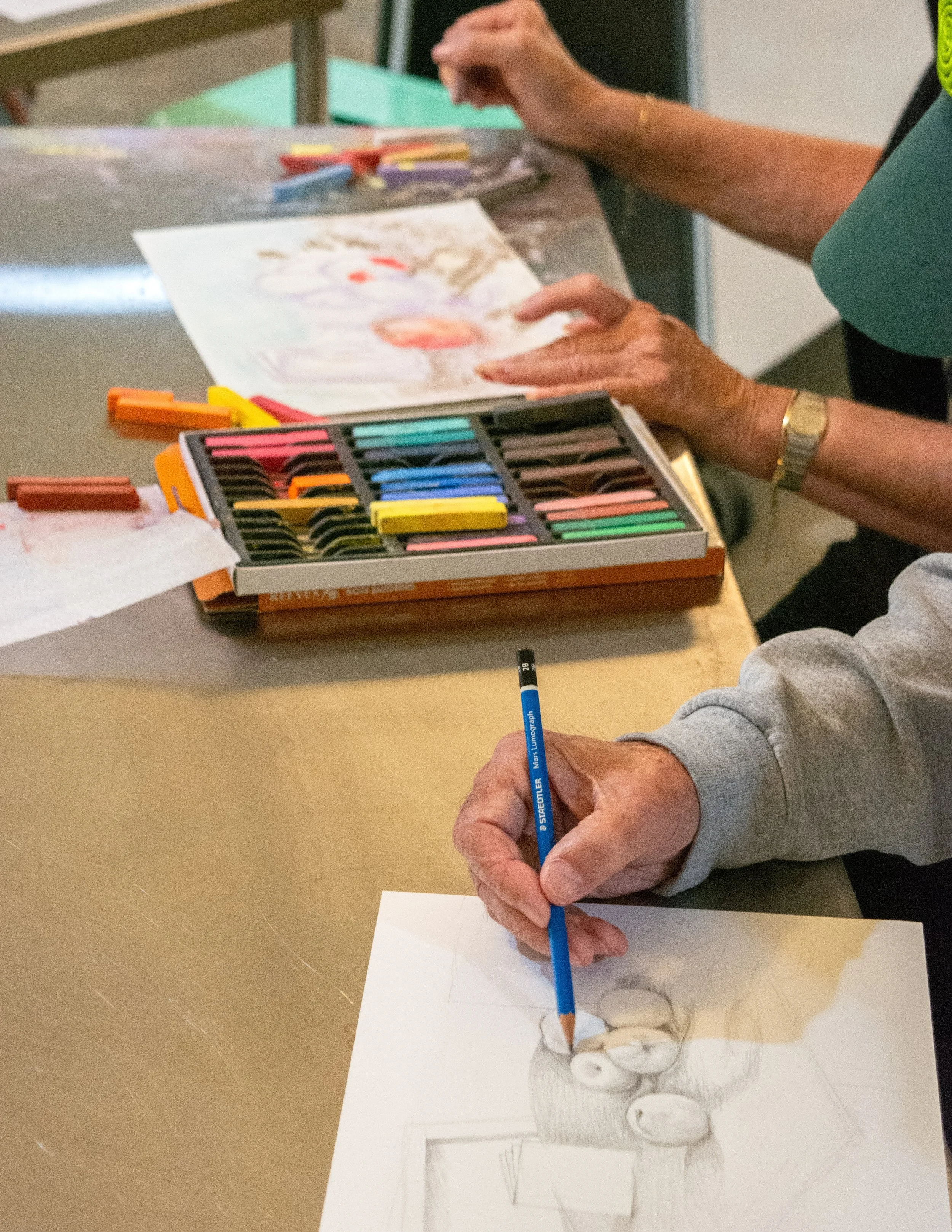 Person drawing a portrait with colored pencils on paper, with a box of pastels and a set of crayons on the table at the Dome House 