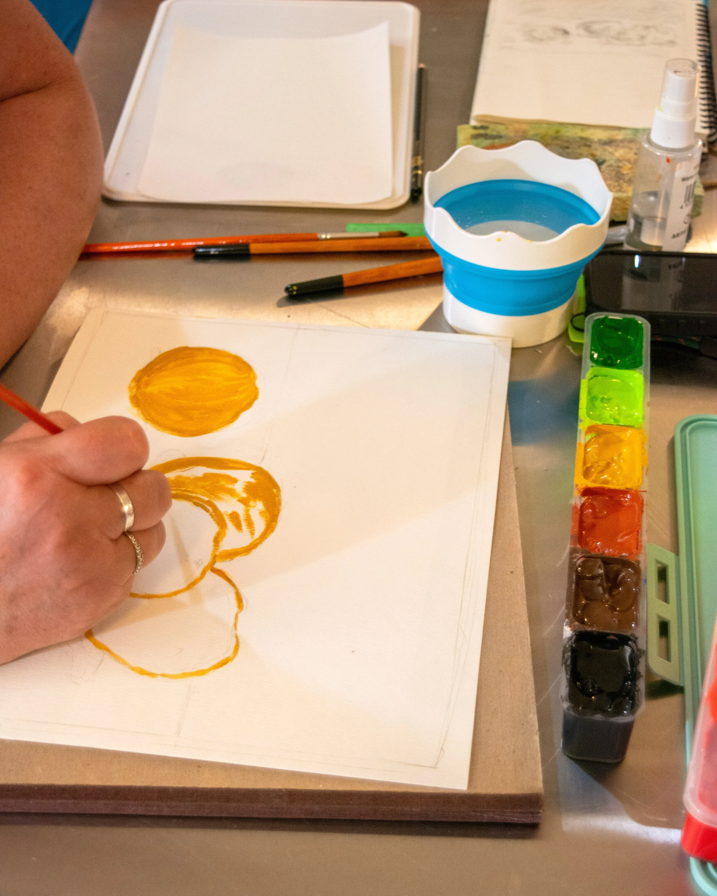 Person painting yellow and orange circles on paper with watercolor paints and brushes, surrounded by paint supplies and stationery.