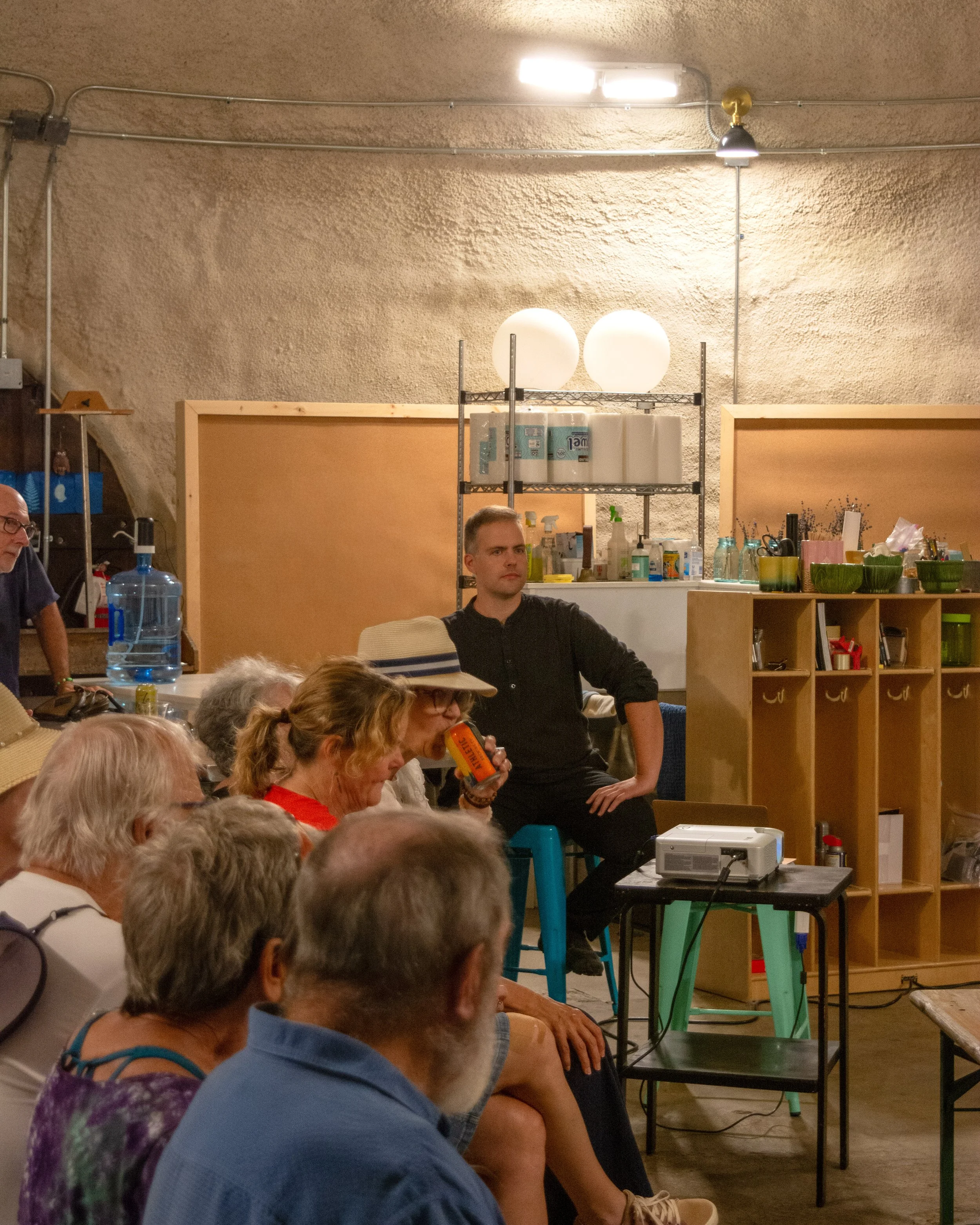 A group of people seated indoors, listening to a presentation. A woman in a hat drinks from a small carton, with a projector on a table in front of her. The background shows shelves with various items and lighting fixtures on the ceiling.