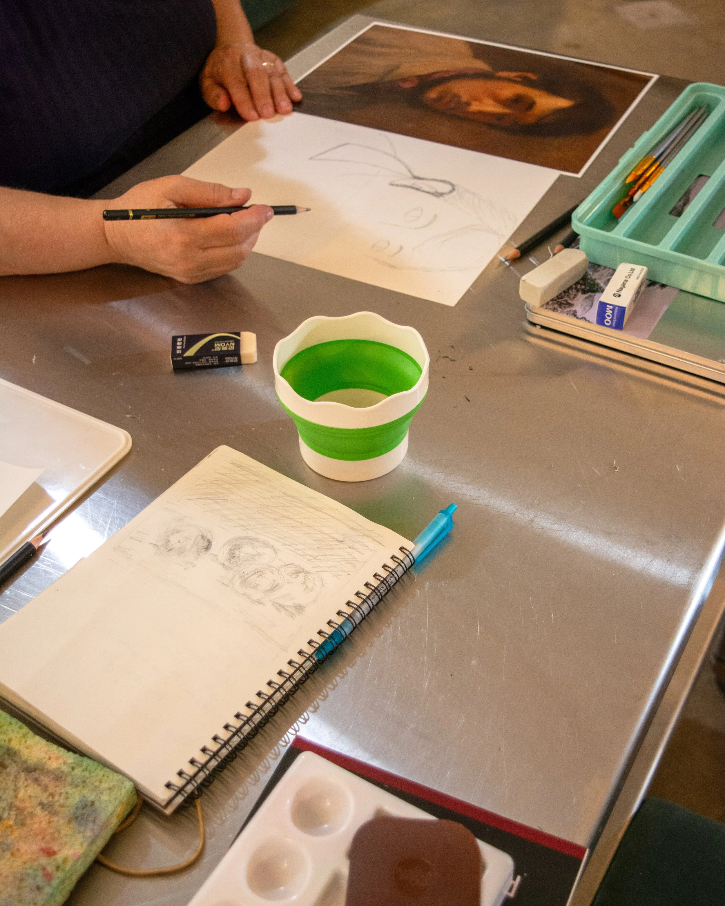 A person drawing sketch of a woman on paper at a workspace with art supplies, including a sketchbook, pencil, eraser, watercolor palette, and reference photos.