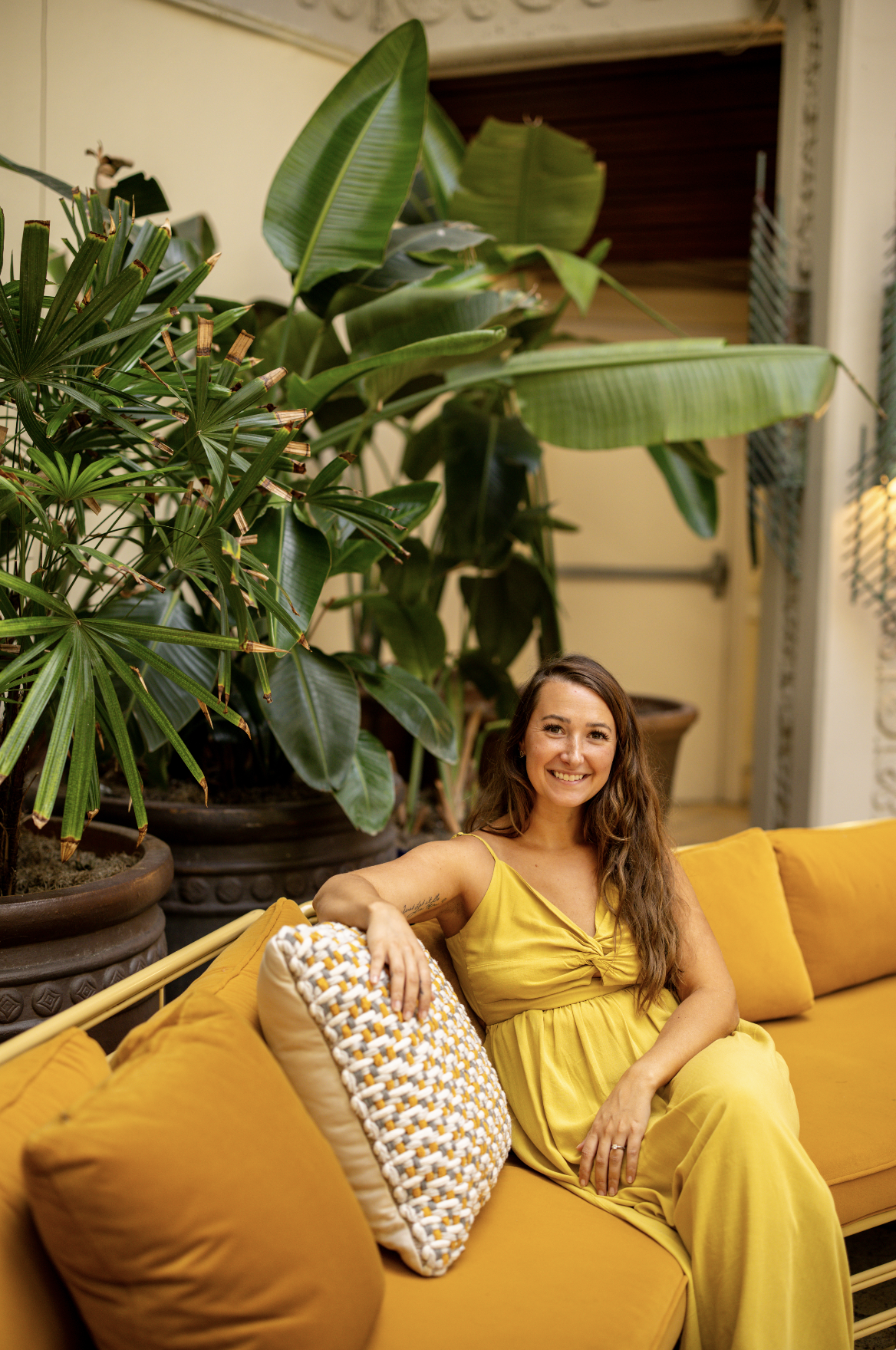 A woman in a yellow dress sitting on a yellow couch, smiling, with large green plants and potted trees behind her.