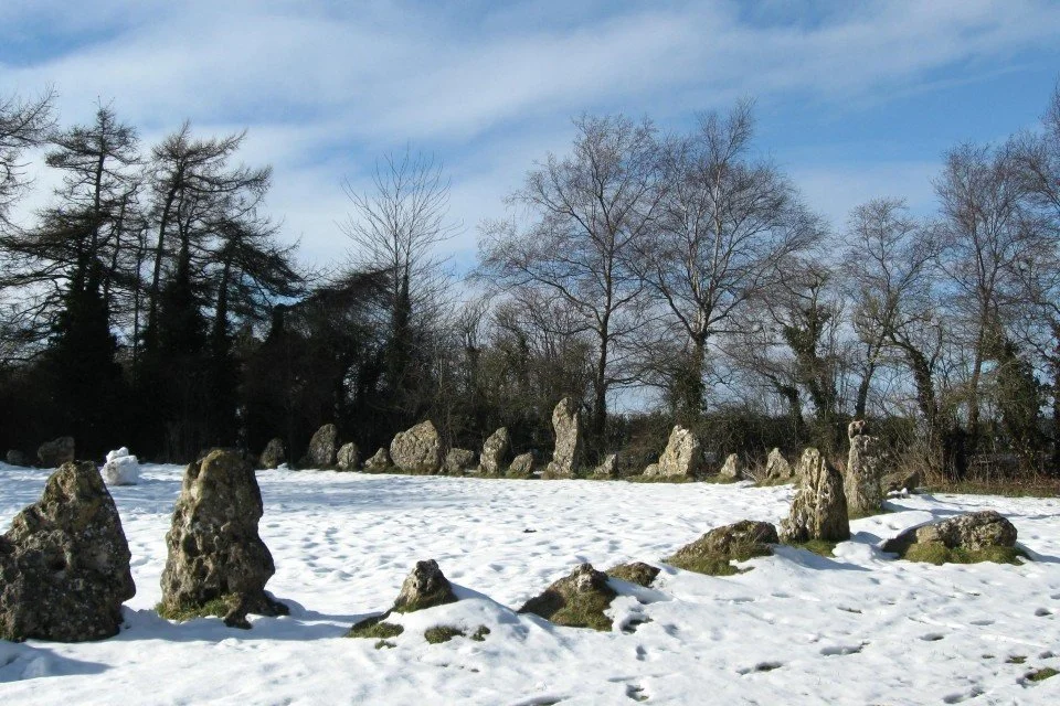 Snow-covered field with standing stones arranged in a circle, and leafless trees in the background under a partly cloudy sky.