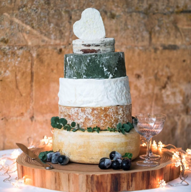 A tiered cheese wheel cake with a rustic wooden base, decorated with greenery and blueberries, topped with a white cheese wedge and a green cheese slice, in front of a brick wall, surrounded by string lights and a pink glass champagne coupe.
