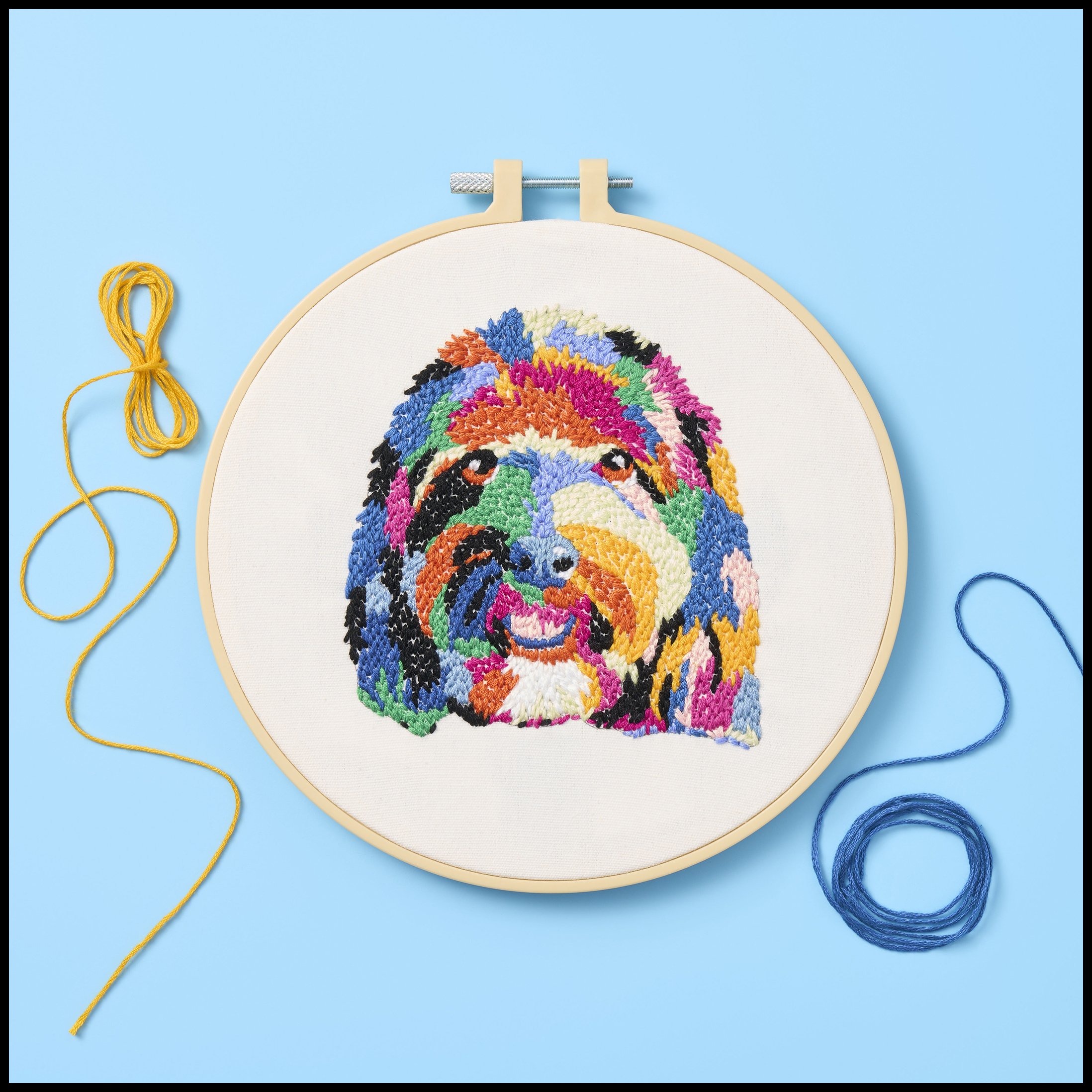 Colorful embroidered portrait of a dog's face in a hoop with yellow and blue threads beside it on a blue background.