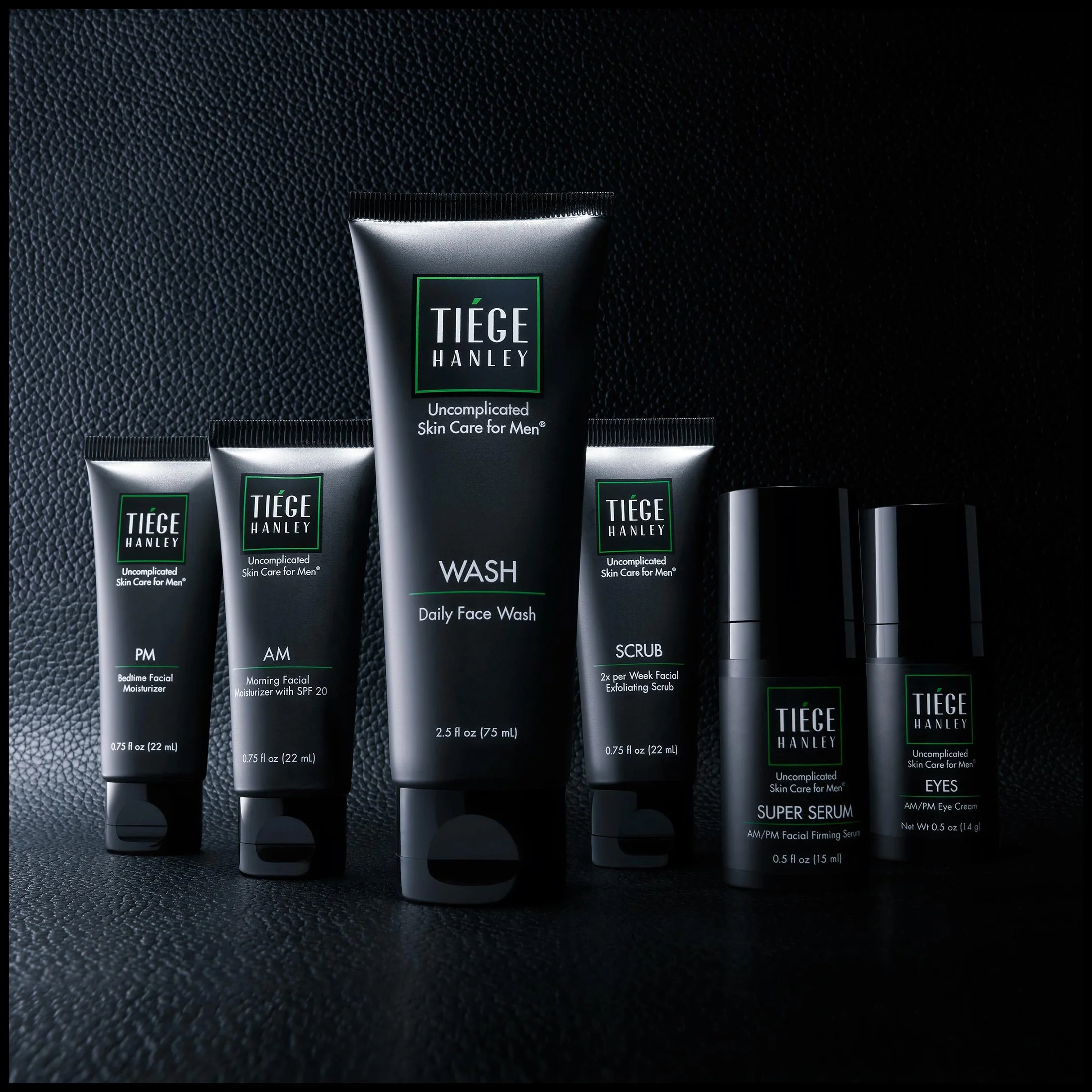 Collection of skincare products from Tiege Hanley, including facial moisturizer, face wash, exfoliating scrub, facial serum, and eye cream, arranged on a black textured surface.  Tiege Hanley.