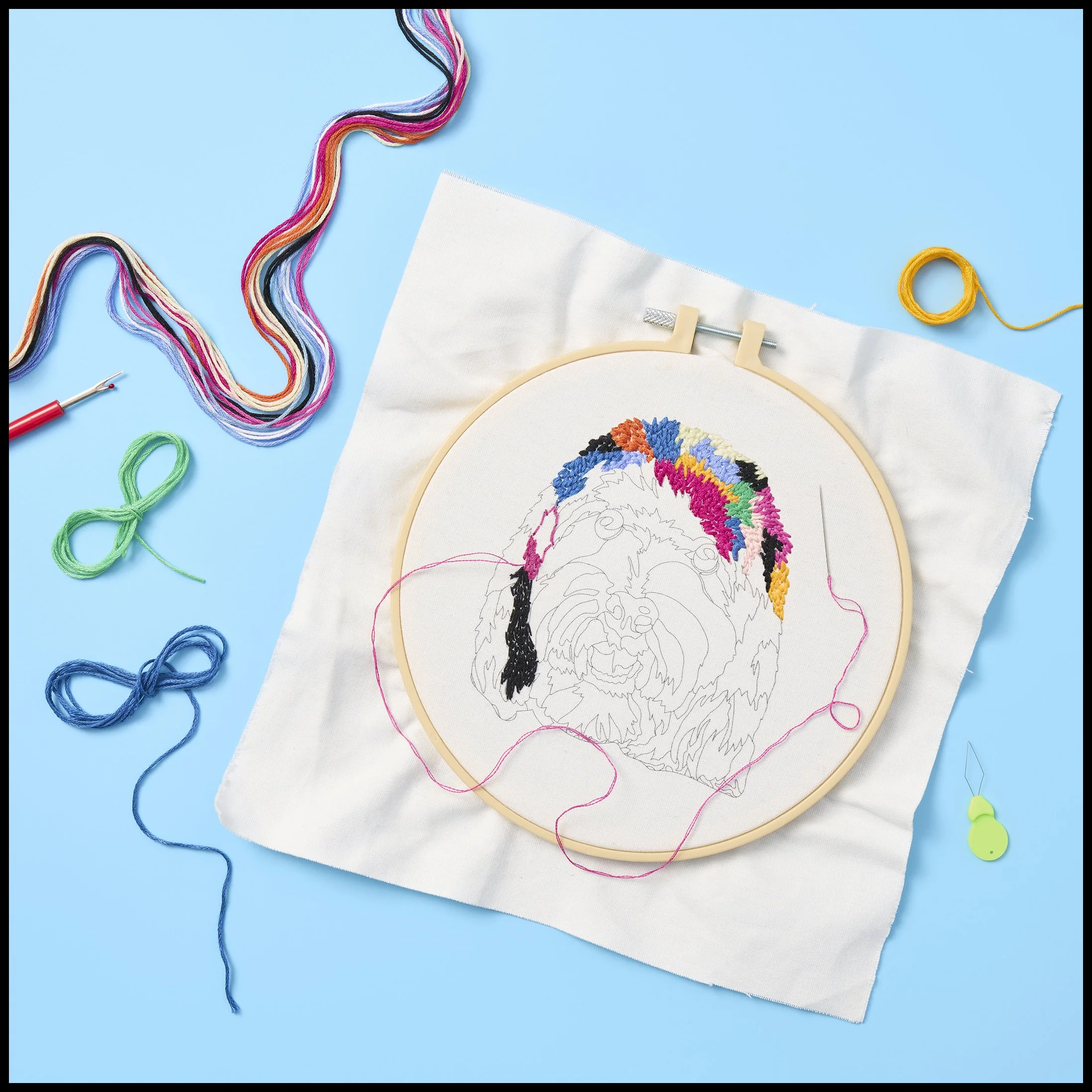 Cross-stitch embroidery of a dog face on fabric with colorful stitches, surrounded by embroidery tools and thread on a blue background.