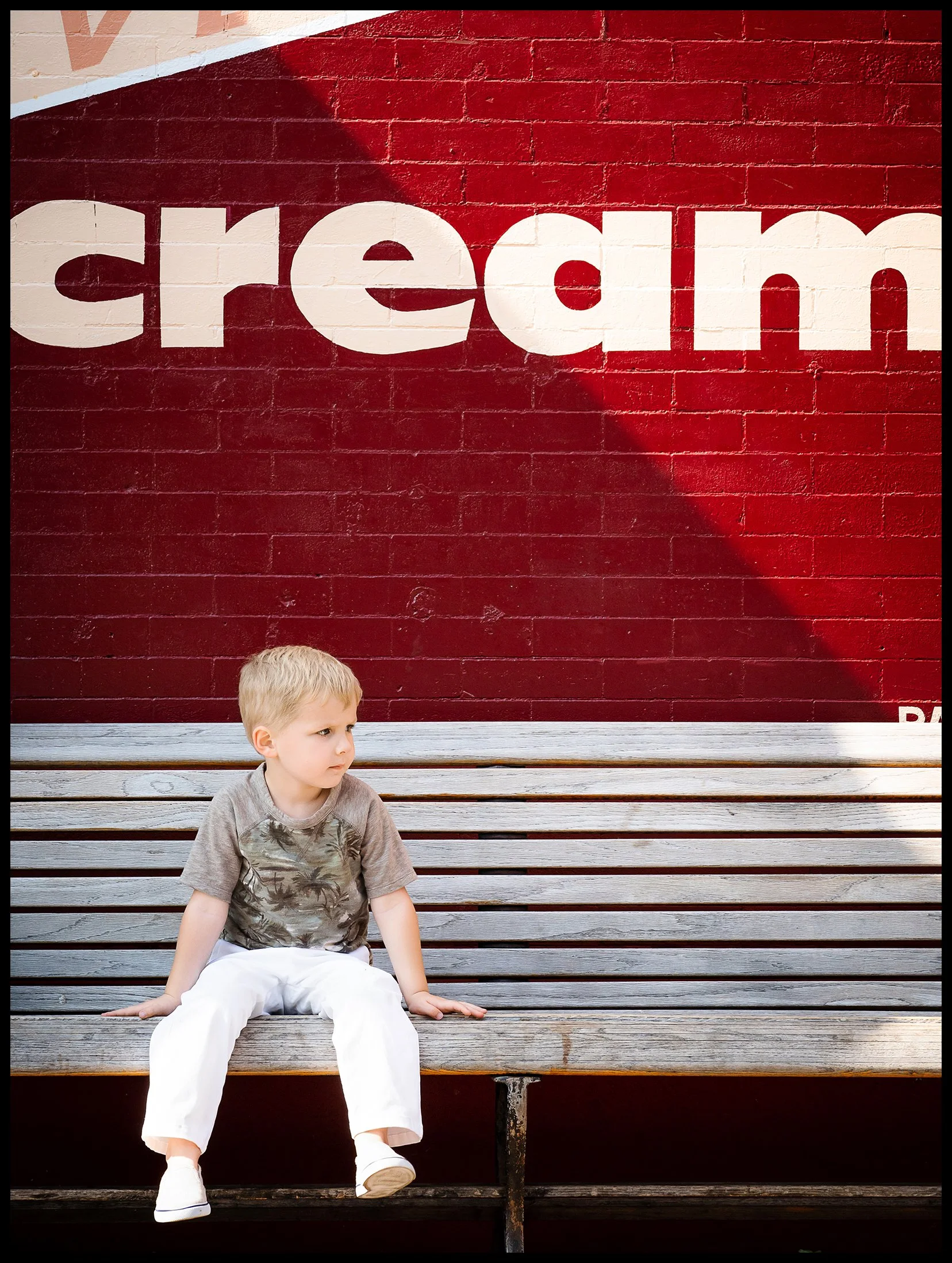 A young boy with blonde hair sitting on a wooden bench in front of a red brick wall with white text.  Minneapolis Kids Lifestyle Advertising Photography Talent Model.