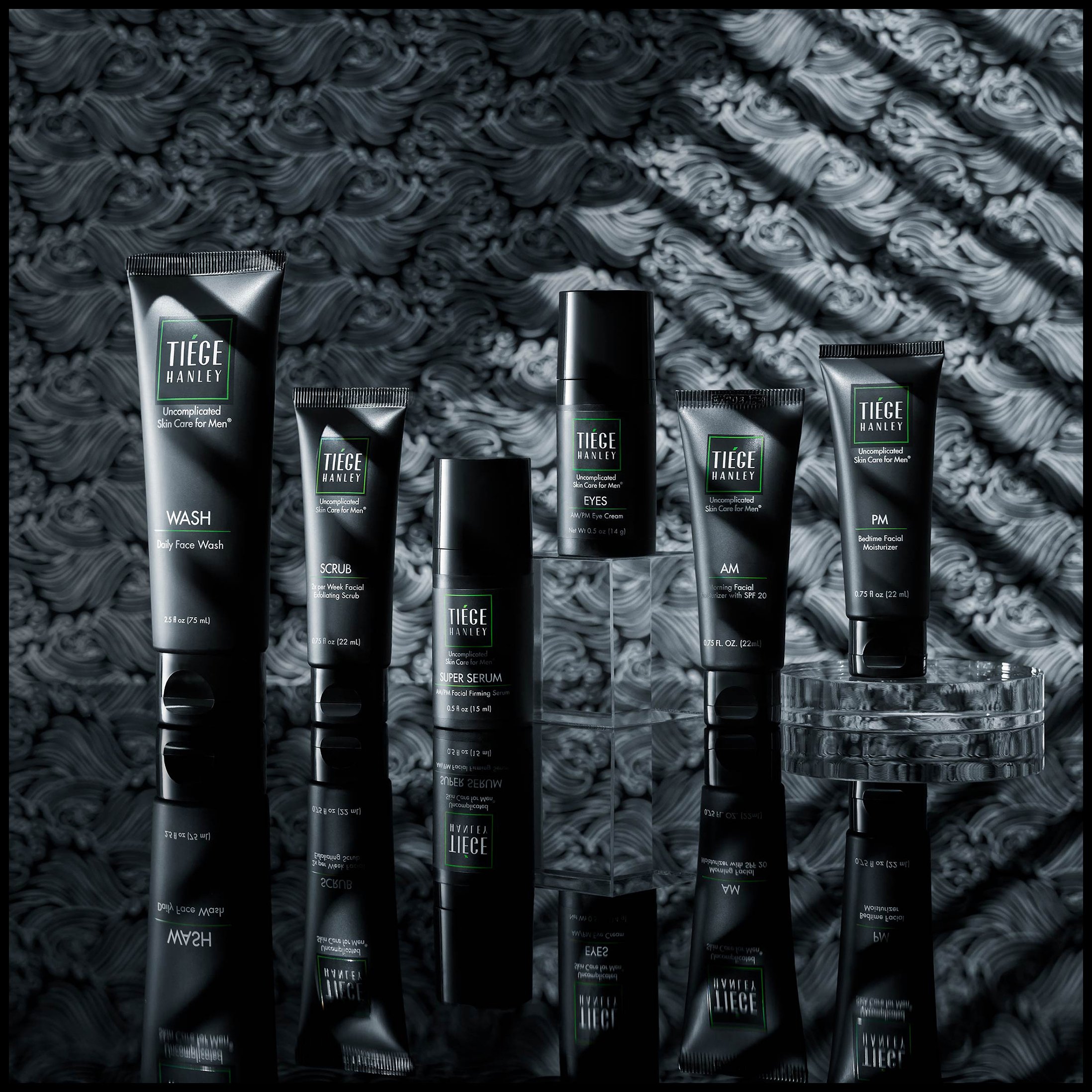 A collection of men's skincare products, including face wash, facial scrub, serum, eye cream, facial moisturizer, and nighttime moisturizer, displayed on a reflective black surface with a textured black and gray background.  Tiege Hanley.