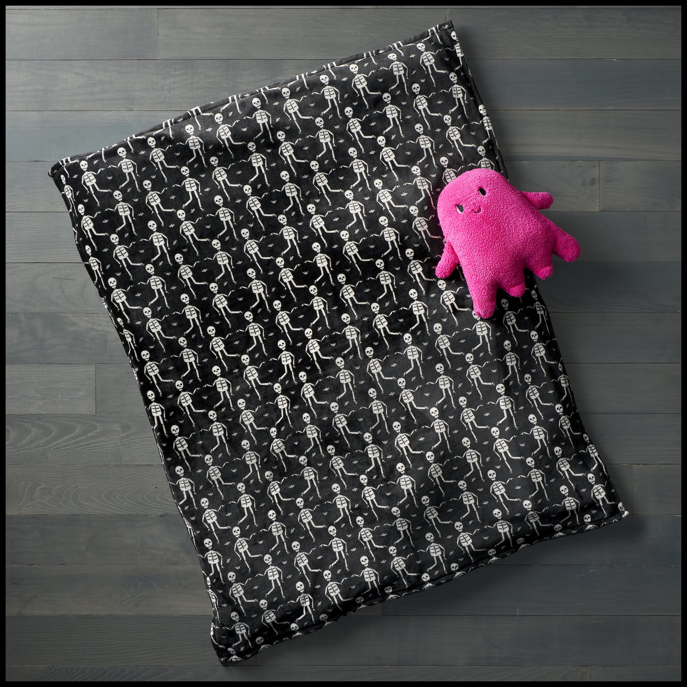 A black patterned fabric with white skeletons and a pink plush octopus toy on a gray wooden floor.  Target HydeAndEek Blanket Printed Skeleton Plush Throw Product Advertising Photography KyleSmithPhoto.