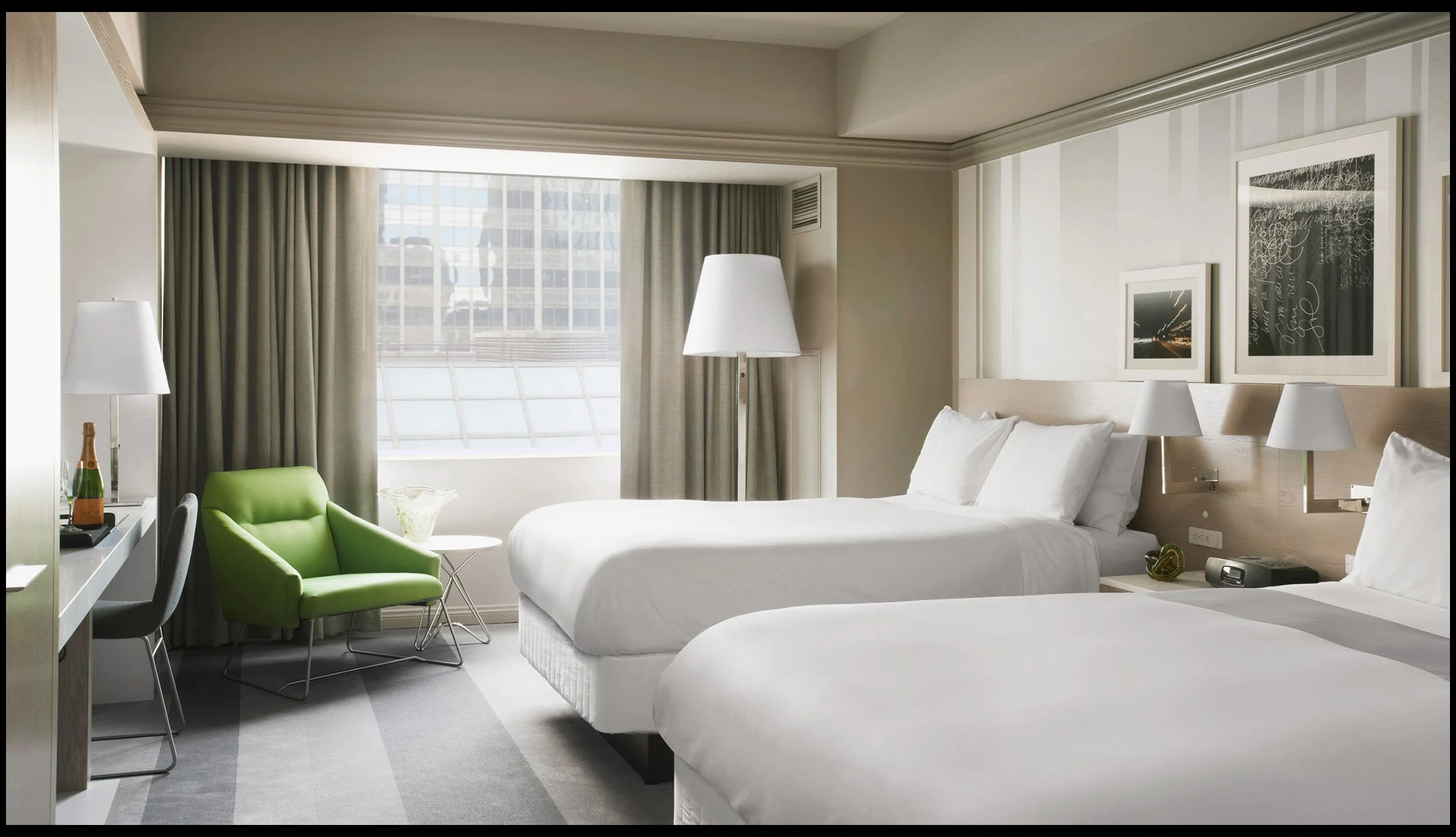 Hotel room with two white beds, a green armchair, a window with city views, and artwork on the wall.