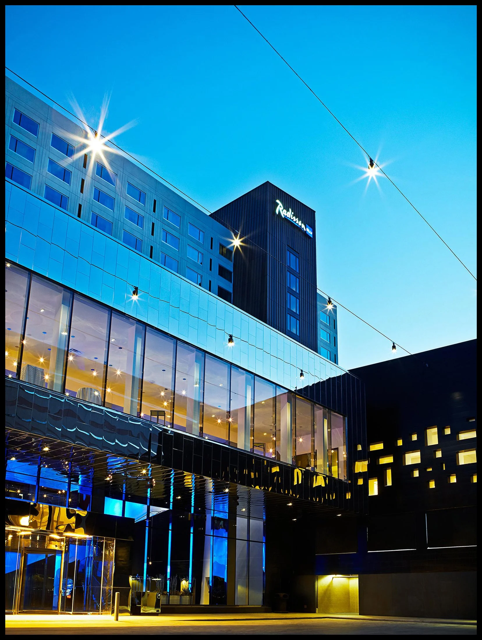 Exterior view of a modern hotel building at dusk with illuminated windows and the "Radisson" sign on the top, featuring glass and black panel architecture, street lighting, and a clear blue sky.