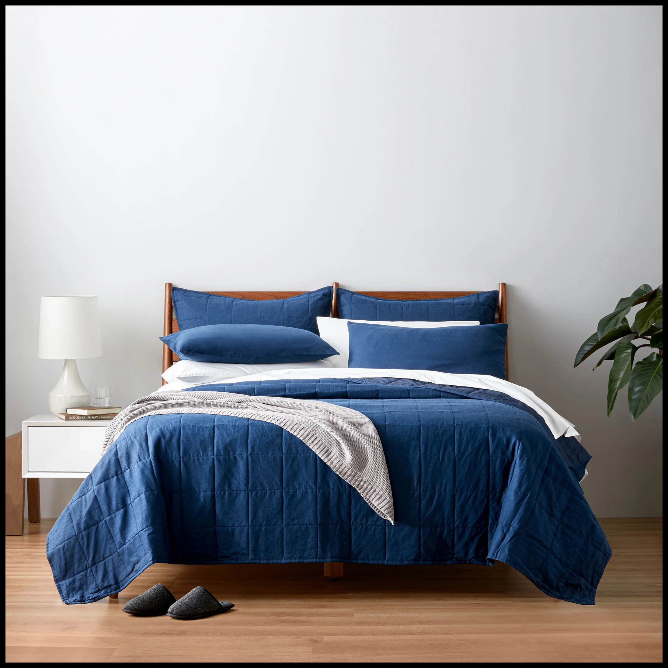 A neatly made bed with a blue quilt, multiple pillows in blue and white, a light gray blanket, and slippers on a hardwood floor, next to a white nightstand with a white lamp and a small plant