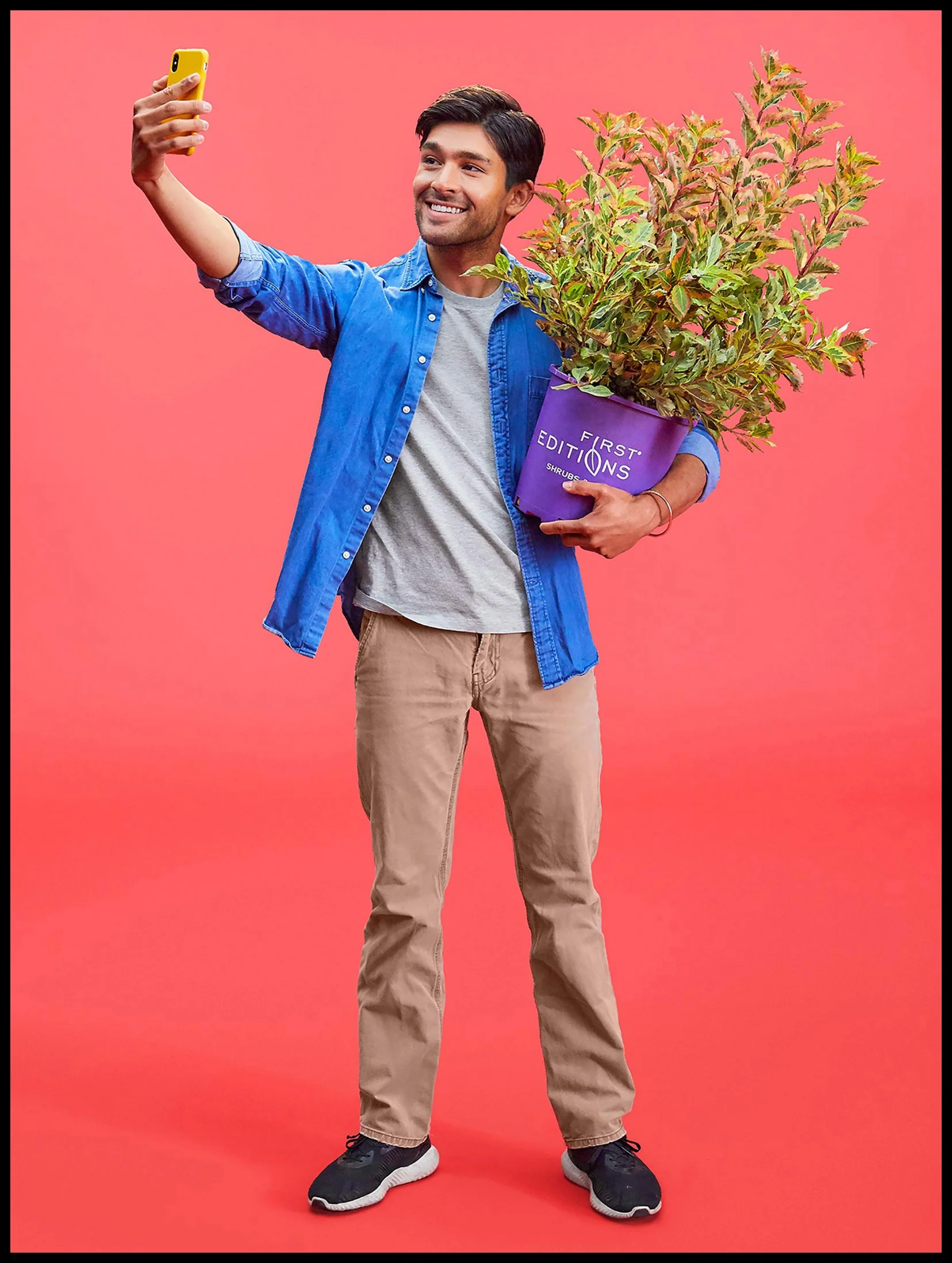 A man holds a yellow phone in one hand and takes a selfie, while holding a purple pot with a large plant in the other hand, standing against a pink background.  BaileyNursery Plants Product Advertising Photography KyleSmithPhoto.