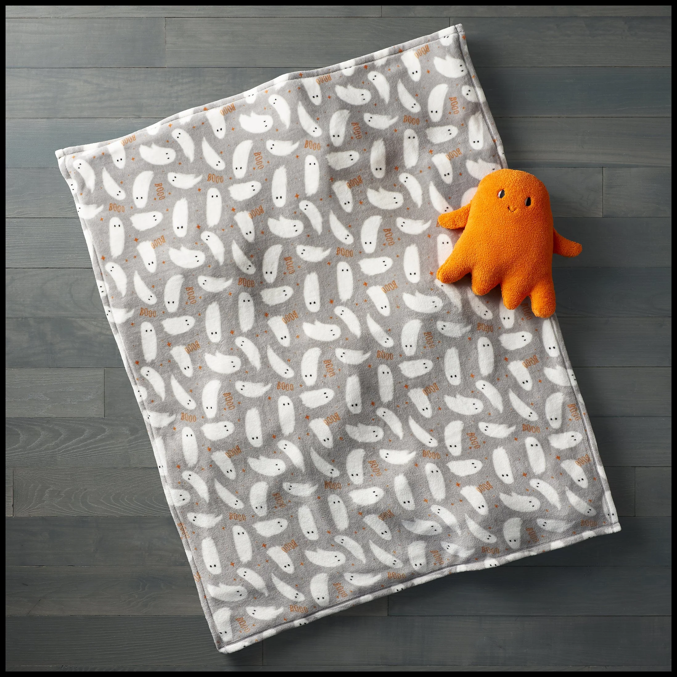 A gray blanket with a pattern of white ducks and the word 'BOO' in orange, laid on a wooden floor, with an orange plush toy shaped like a ghost on top.  Target HydeAndEek Blanket Print Ghost Plush Throw Product Advertising Photography KyleSmithPhoto.