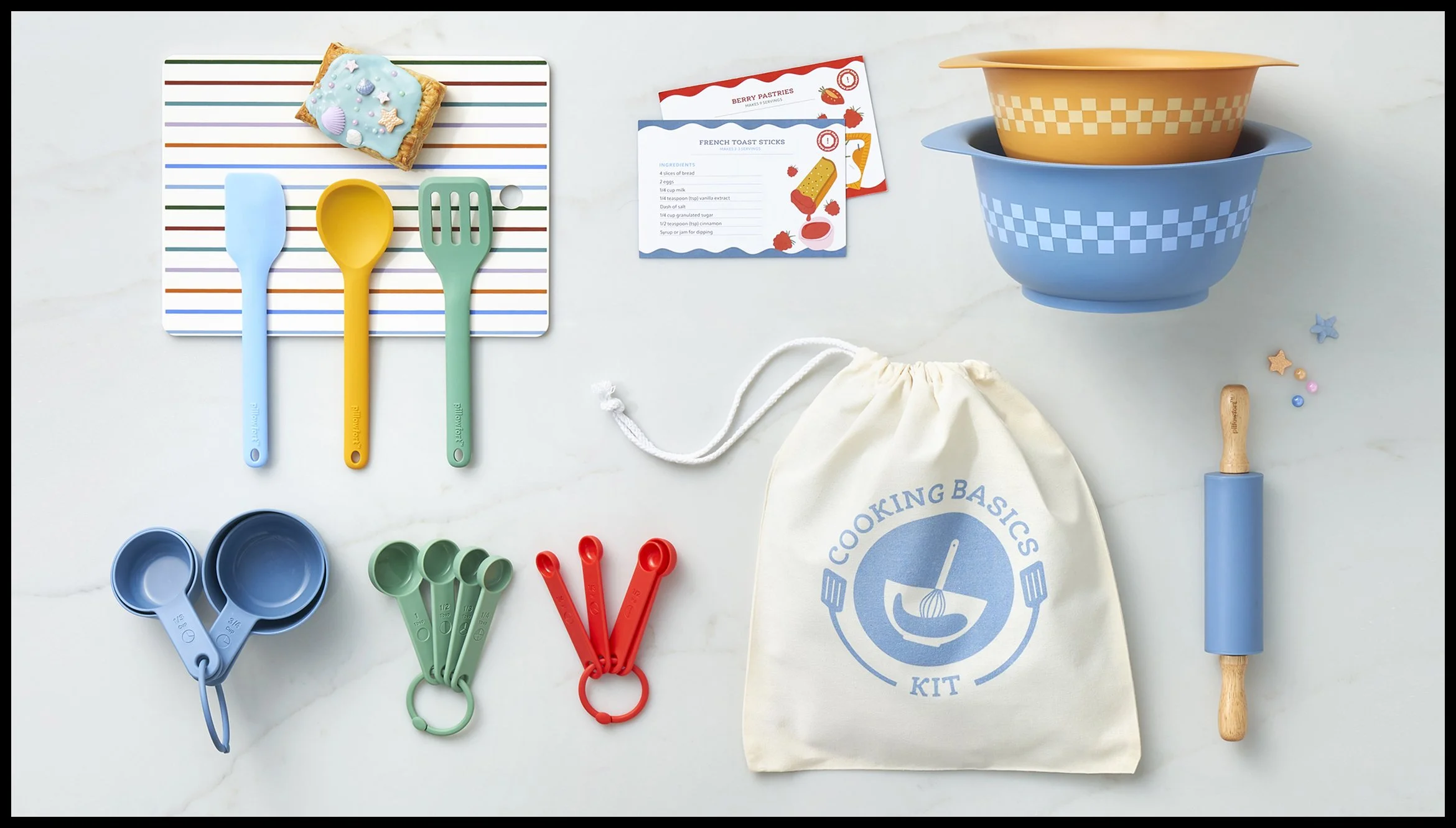 A cooking kit for kids including colorful measuring cups, spoons, a rolling pin, mixing bowls, recipe cards, a cotton bag, and small decorative items on a light surface.