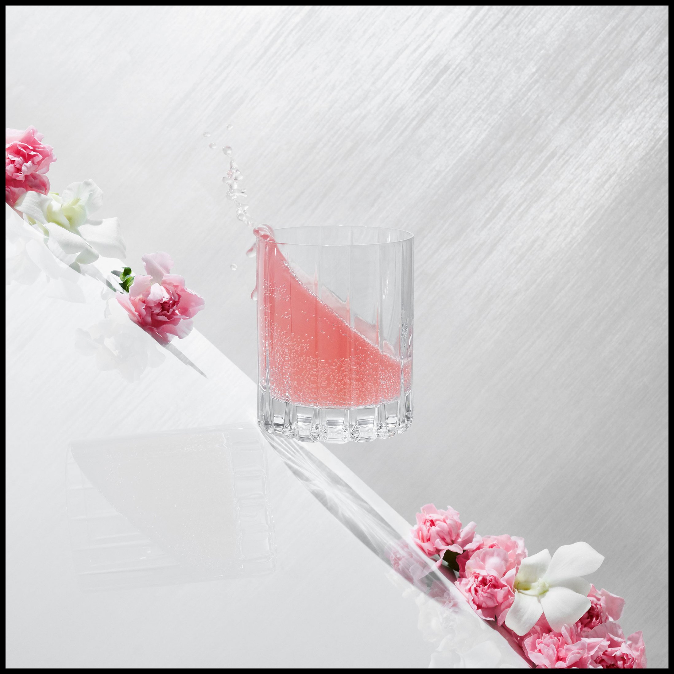 A glass with pink liquid and a slice of pink fruit or vegetable, with water splashing out, surrounded by pink and white flowers on a white surface.  Beverage Drink Drinkware Pink Flowers Product Advertising Photography KyleSmithPhoto.