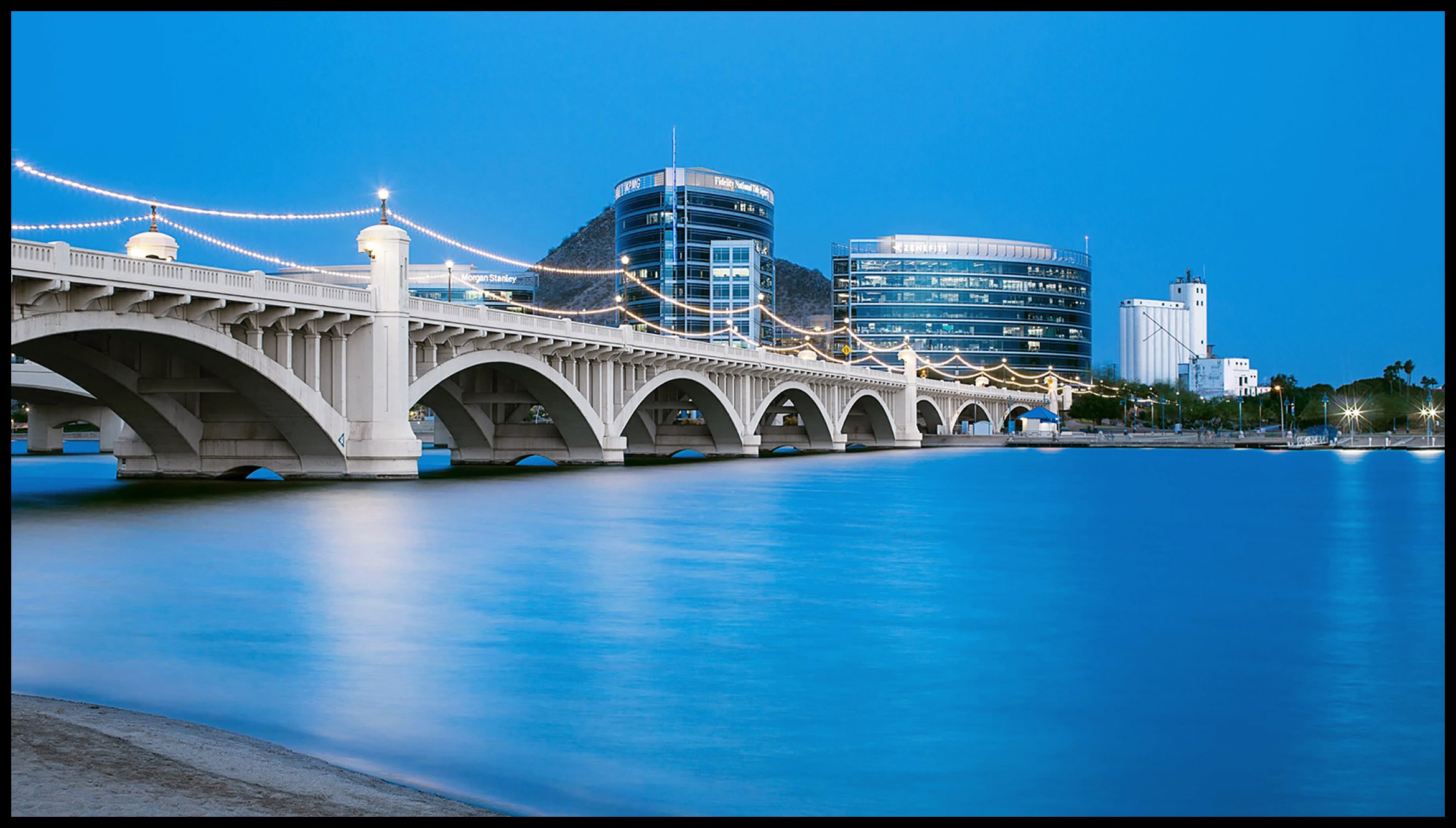 Night view of a bridge over a body of water with a city skyline featuring modern buildings and lights in the background.