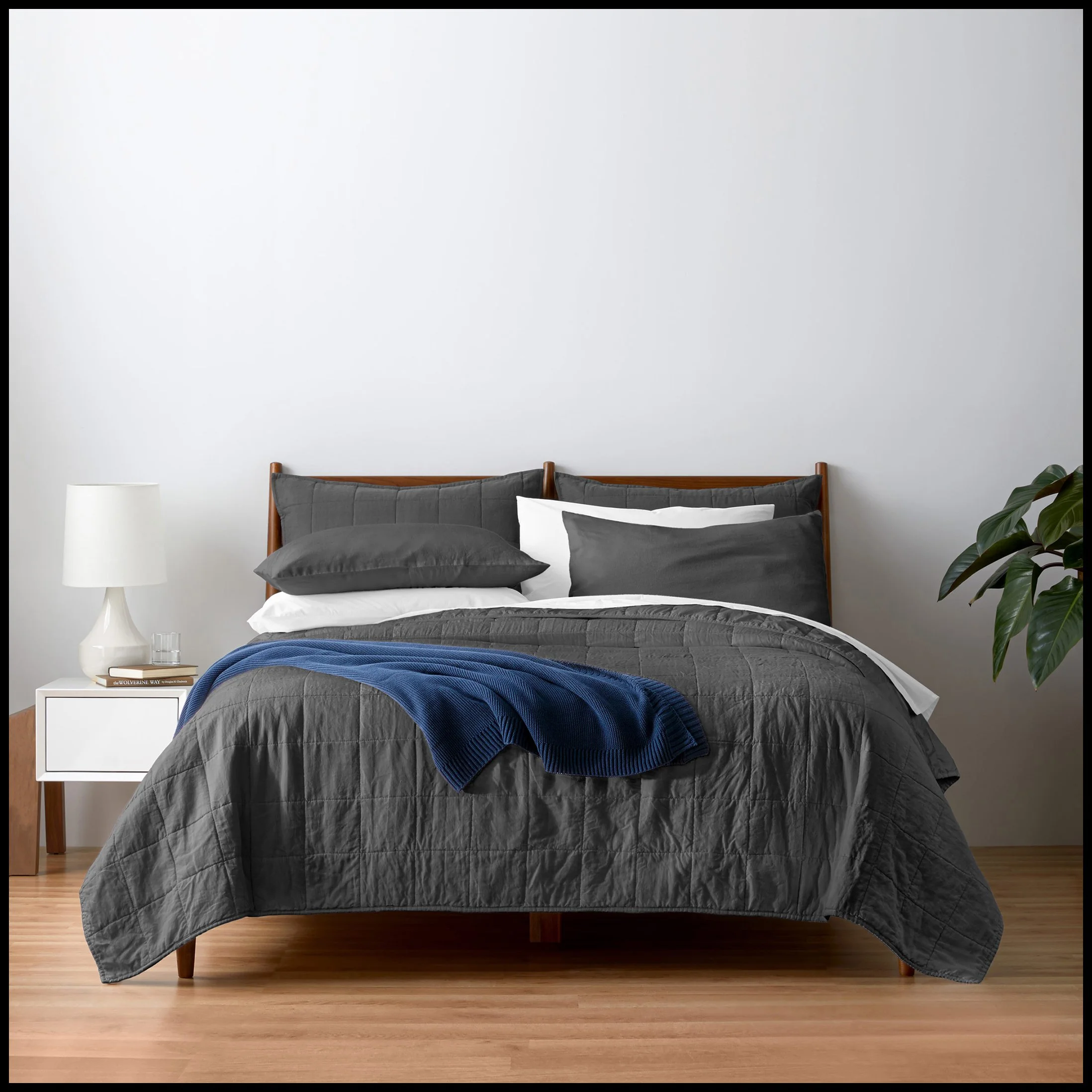 A bedroom with a bed dressed in gray bedding, black and white pillows, a blue throw blanket, a white nightstand, a white table lamp, a small glass, a book, and a green potted plant in the corner, against a plain white wall and wooden flooring.