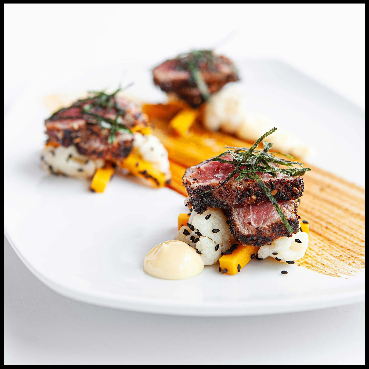 Close-up of a gourmet appetizer with grilled beef, cauliflower, and yellow peppers garnished with sliced seaweed, with sauce and grill marks on a white plate.