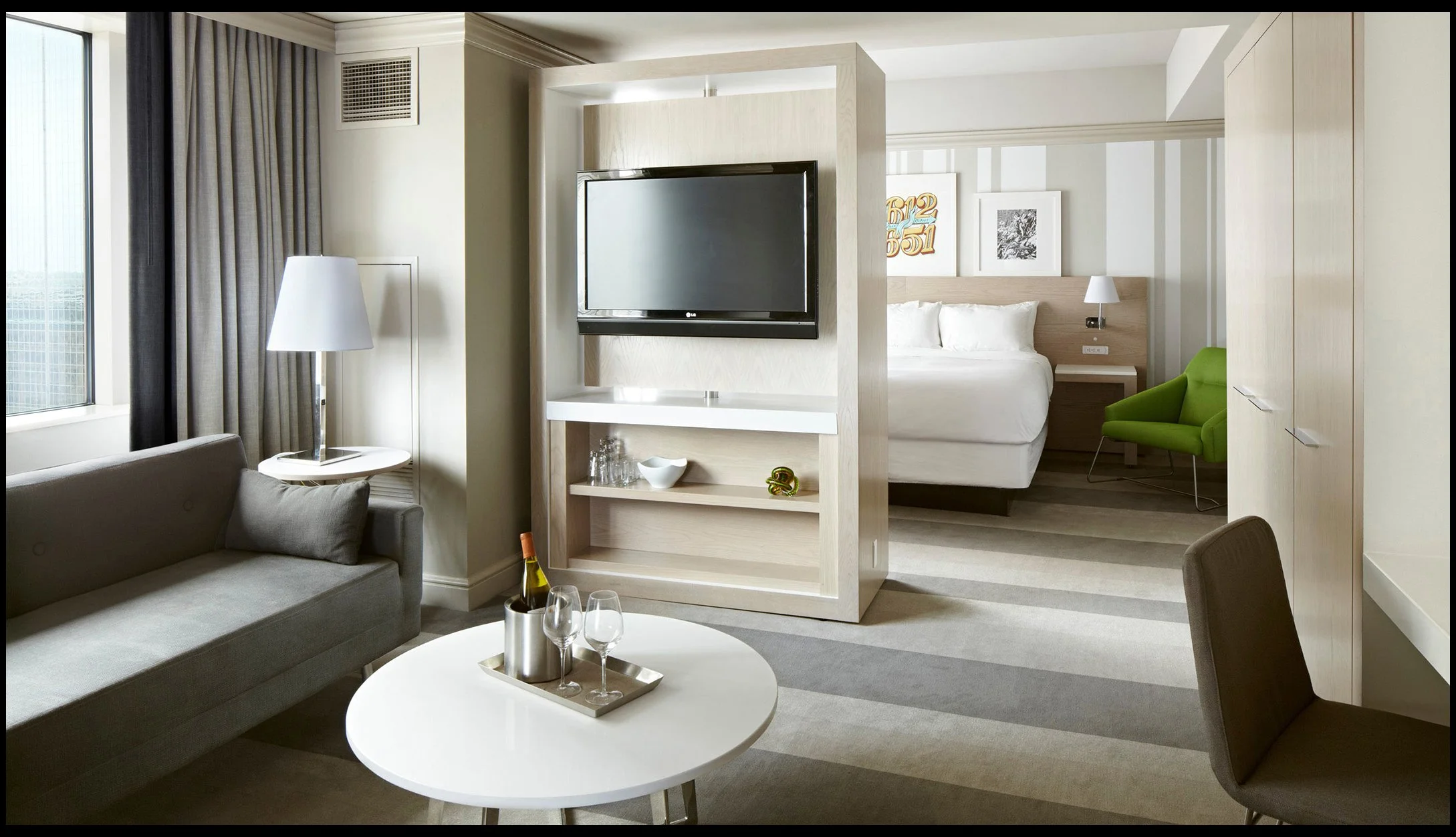 Modern hotel suite featuring a living area with a gray sofa, white coffee table with wine and glasses, and a standing lamp beside windows. The room has a divider with a mounted TV, opening to a bedroom with a white bed, nightstand, and a green accent