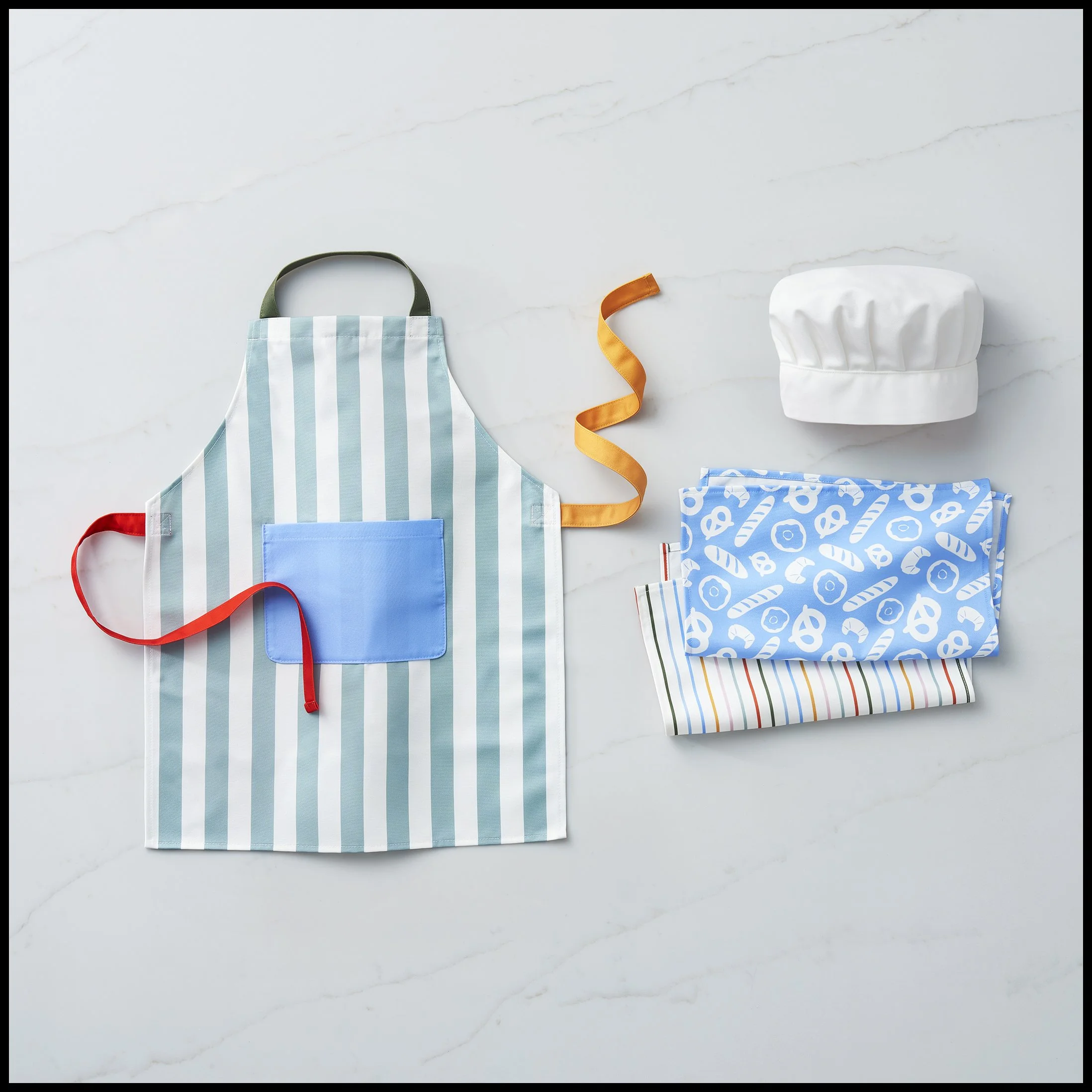 A striped apron with a blue pocket, a white chef's hat, and various colorful kitchen towels and ribbons on a white surface.