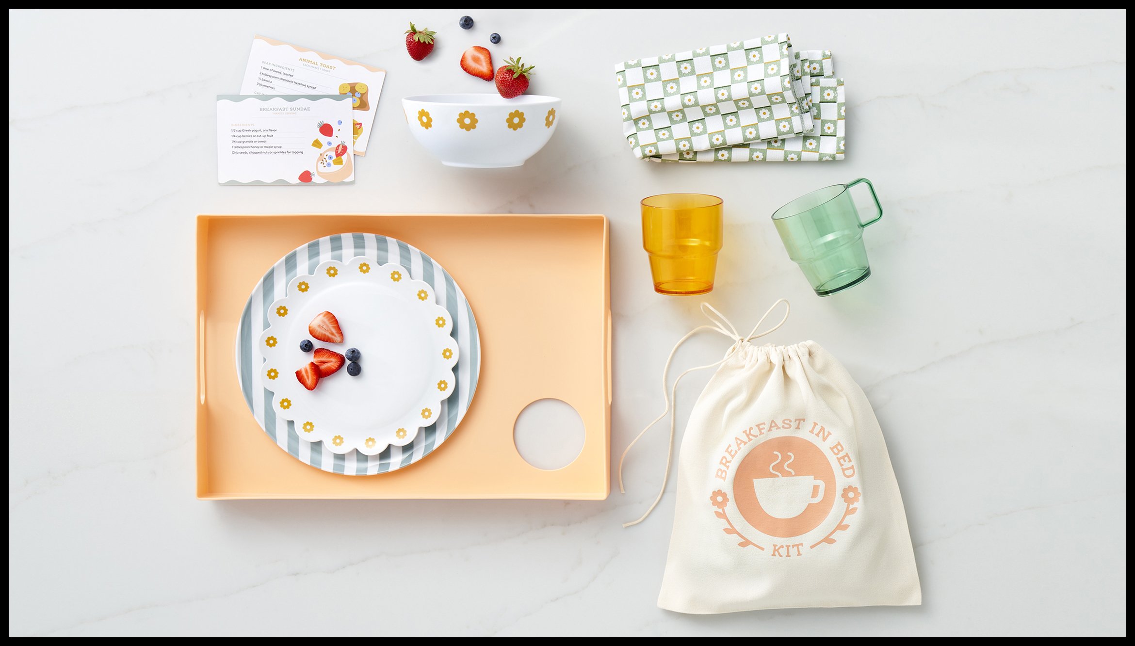 A breakfast in bed kit with a white plate of desserts topped with strawberries and blueberries, two colorful glasses, a bowl with strawberries, blueberries, and a strawberry plant, folded napkins with a green floral pattern, recipe cards, and a cloth