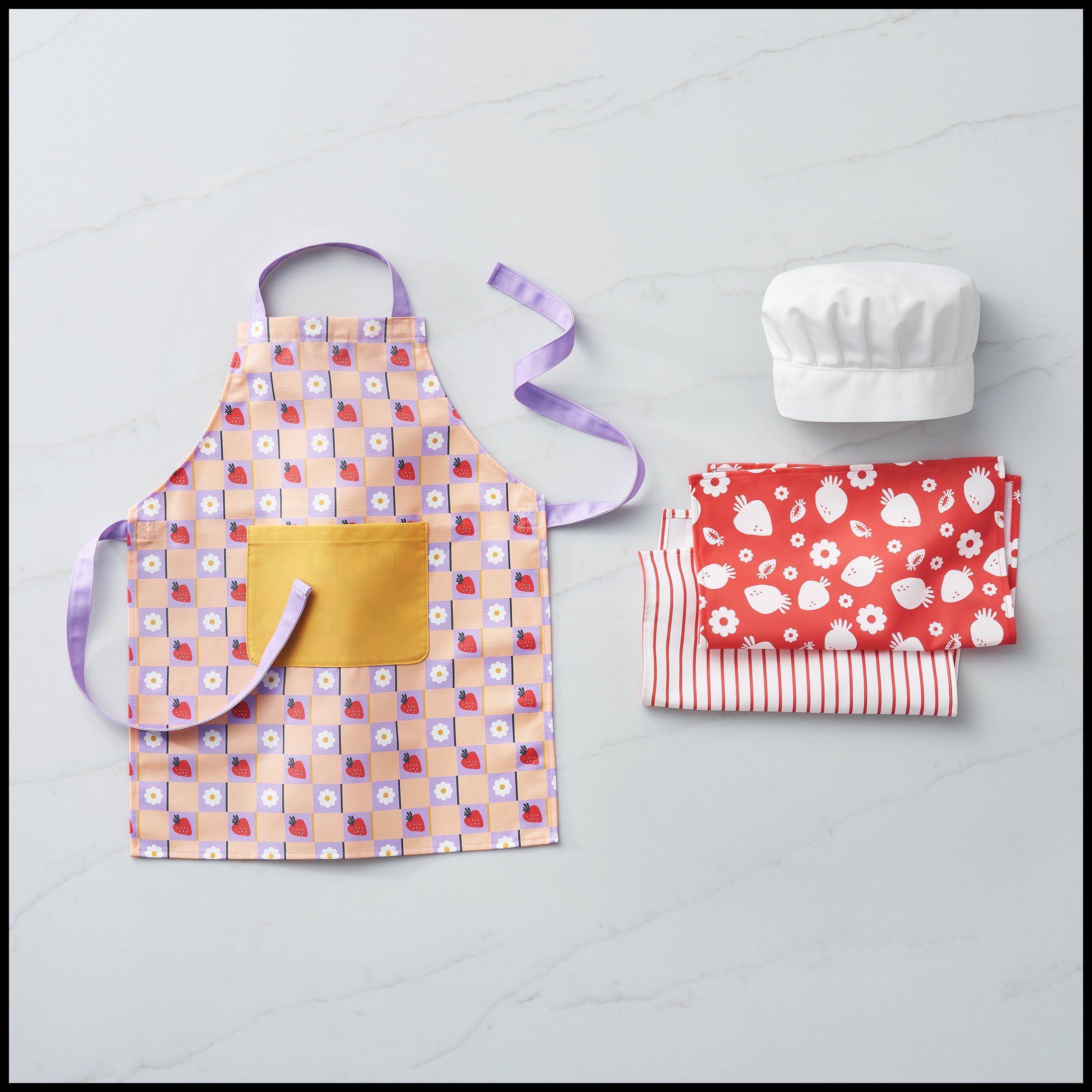 A colorful children's apron with strawberry and daisies pattern, a yellow pocket, and purple ties, placed on a marble surface along with folded red and white patterns, a white chef's hat, and red and white striped fabric.