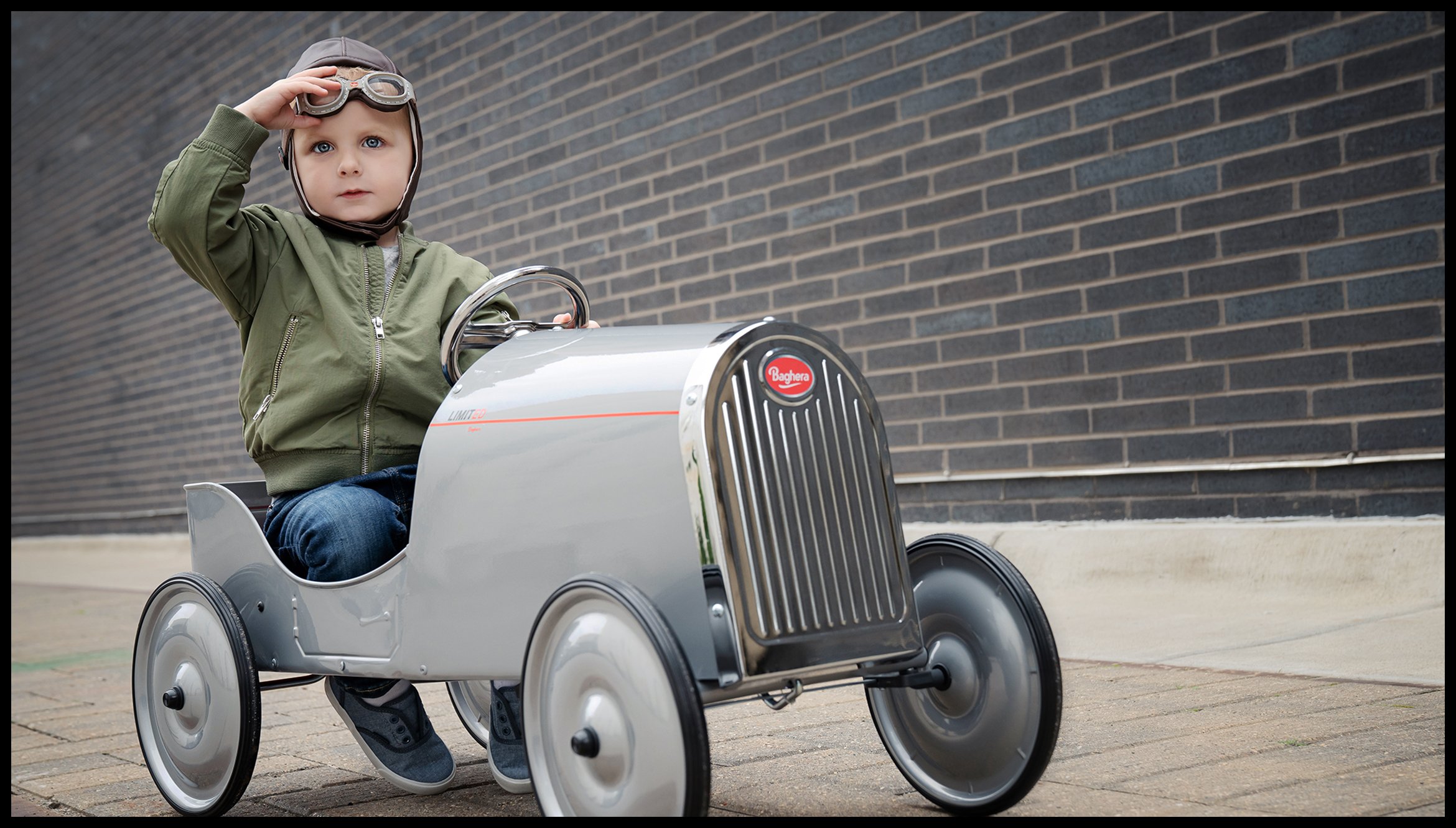 A young child sitting in a vintage silver toy car, wearing a green jacket, blue jeans, gray shoes, and goggles, saluting with one hand.  Minneapolis Kids Lifestyle Advertising Photography Talent Model.
