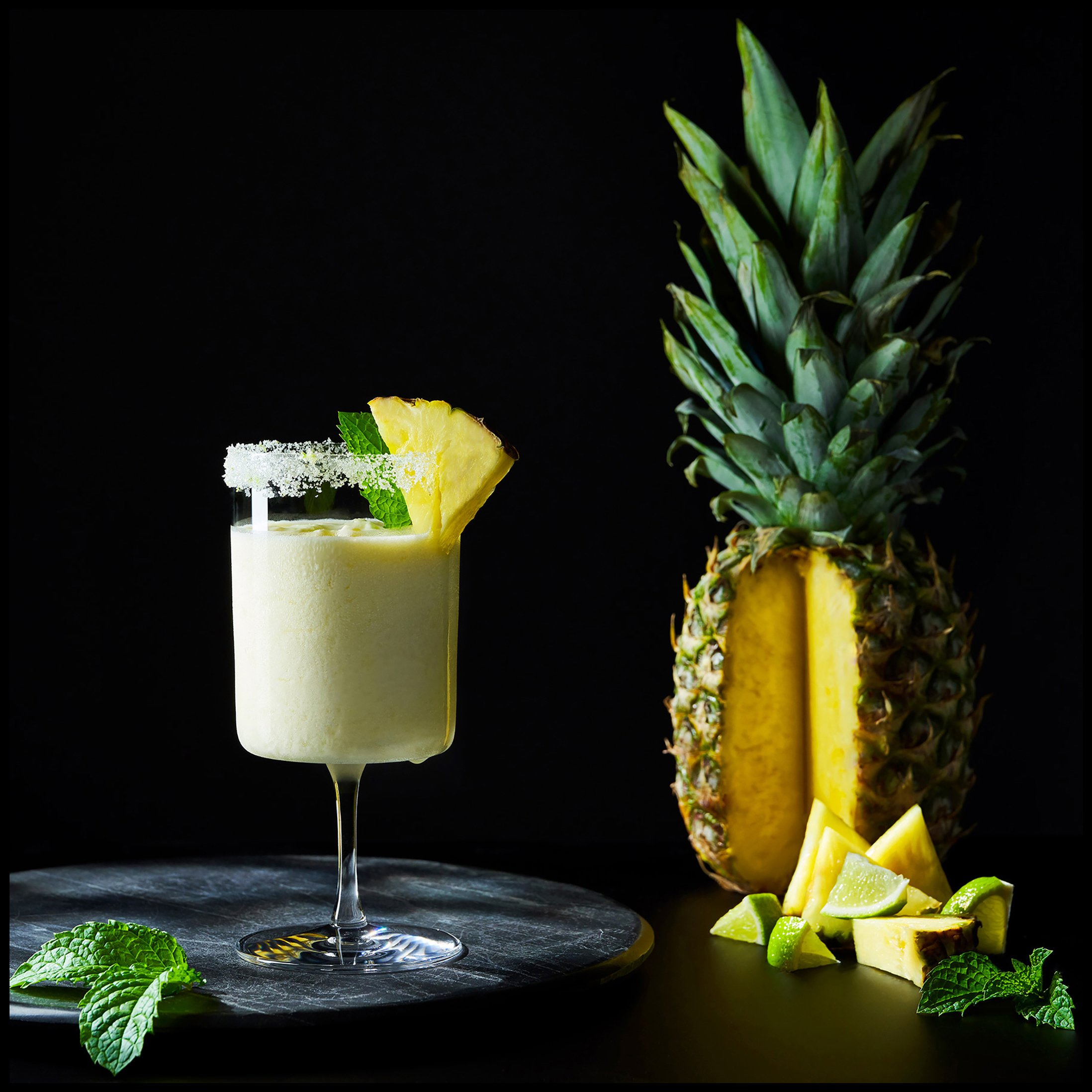 A pineapple cocktail garnished with pineapple wedge and mint leaves, next to a whole pineapple with a cut section and small pineapple pieces surrounded by lime wedges, on a black surface with a dark background.