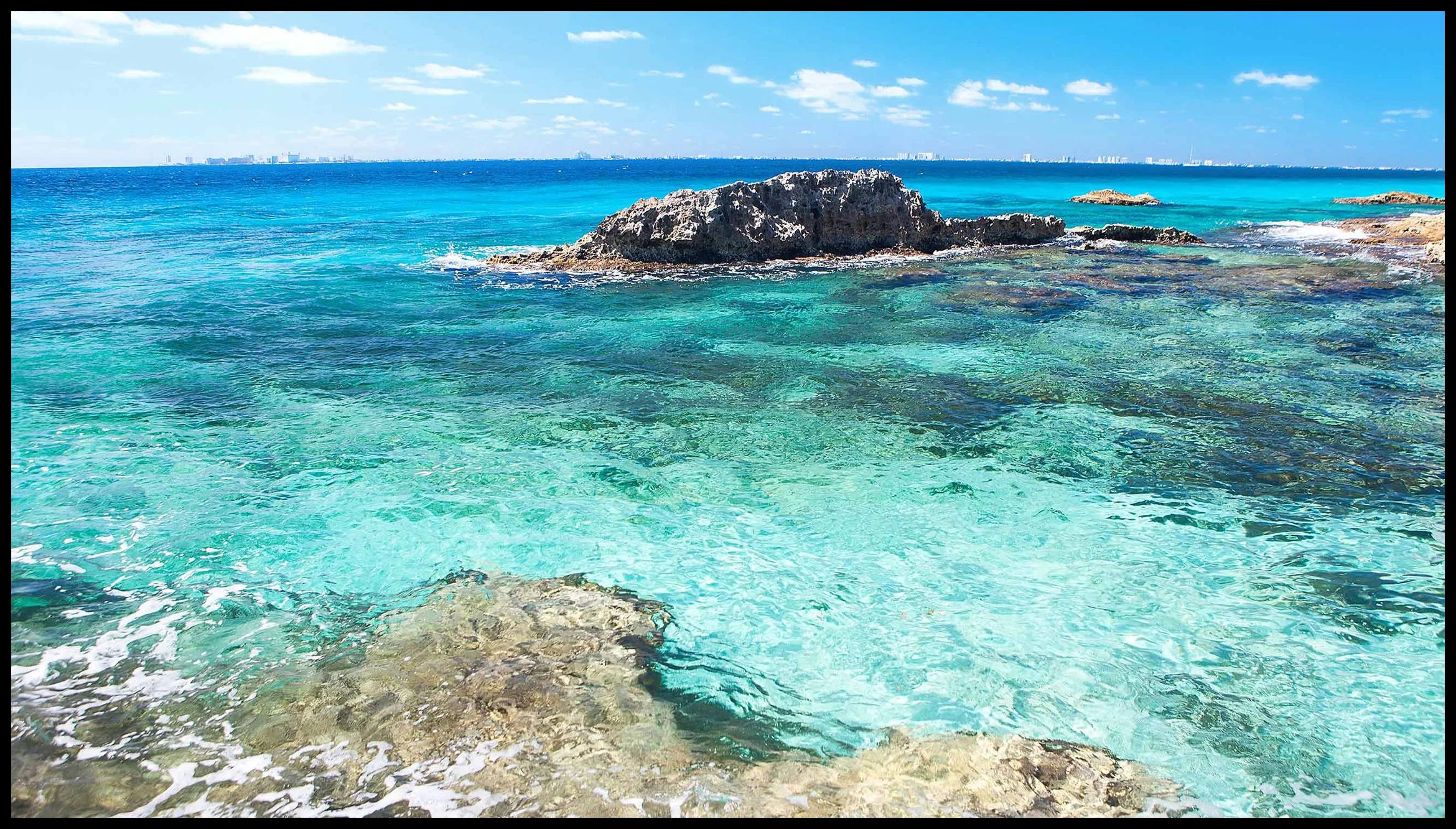 Turquoise ocean water surrounding rocks on a sunny day with a city skyline in the distance under a partly cloudy sky.  Art Prints Isla Mujeres Mexico Island.