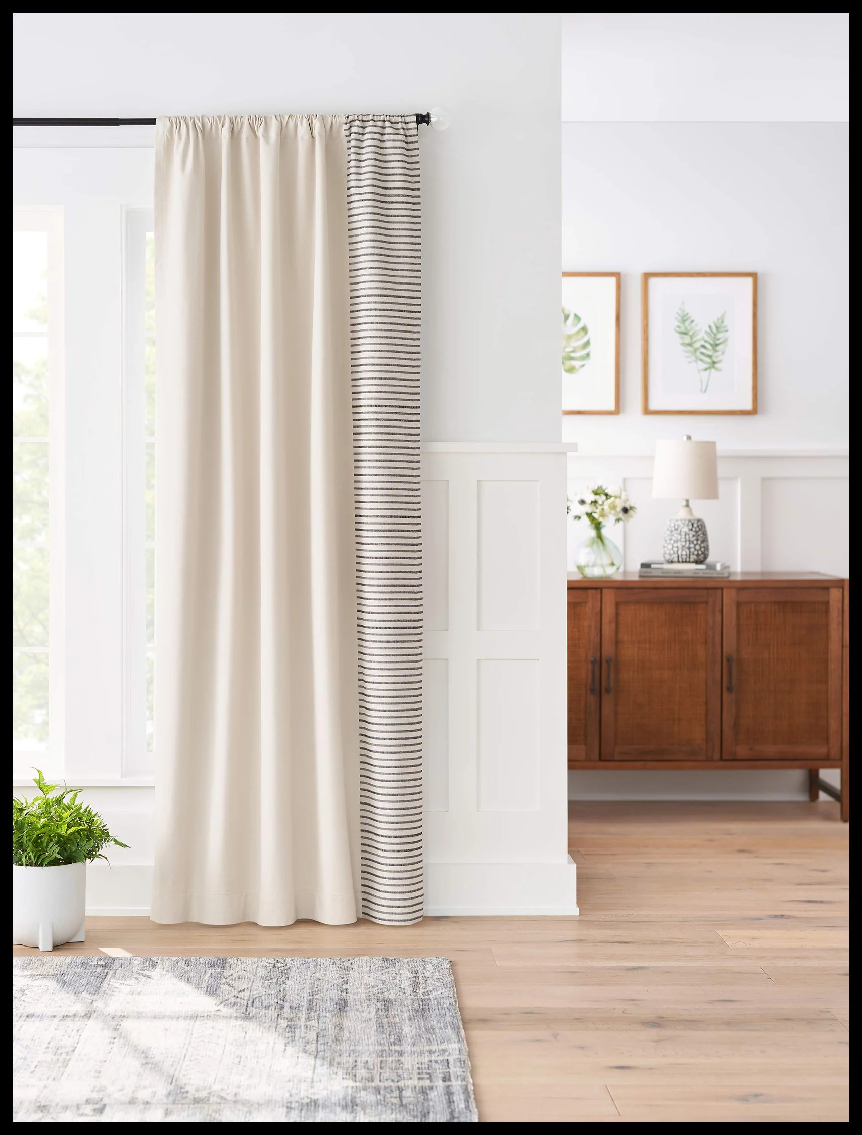 Living room with a window covered by beige and striped curtains, a potted plant on the floor, a wooden sideboard with a lamp and flowers, framed leaf prints on the wall.
