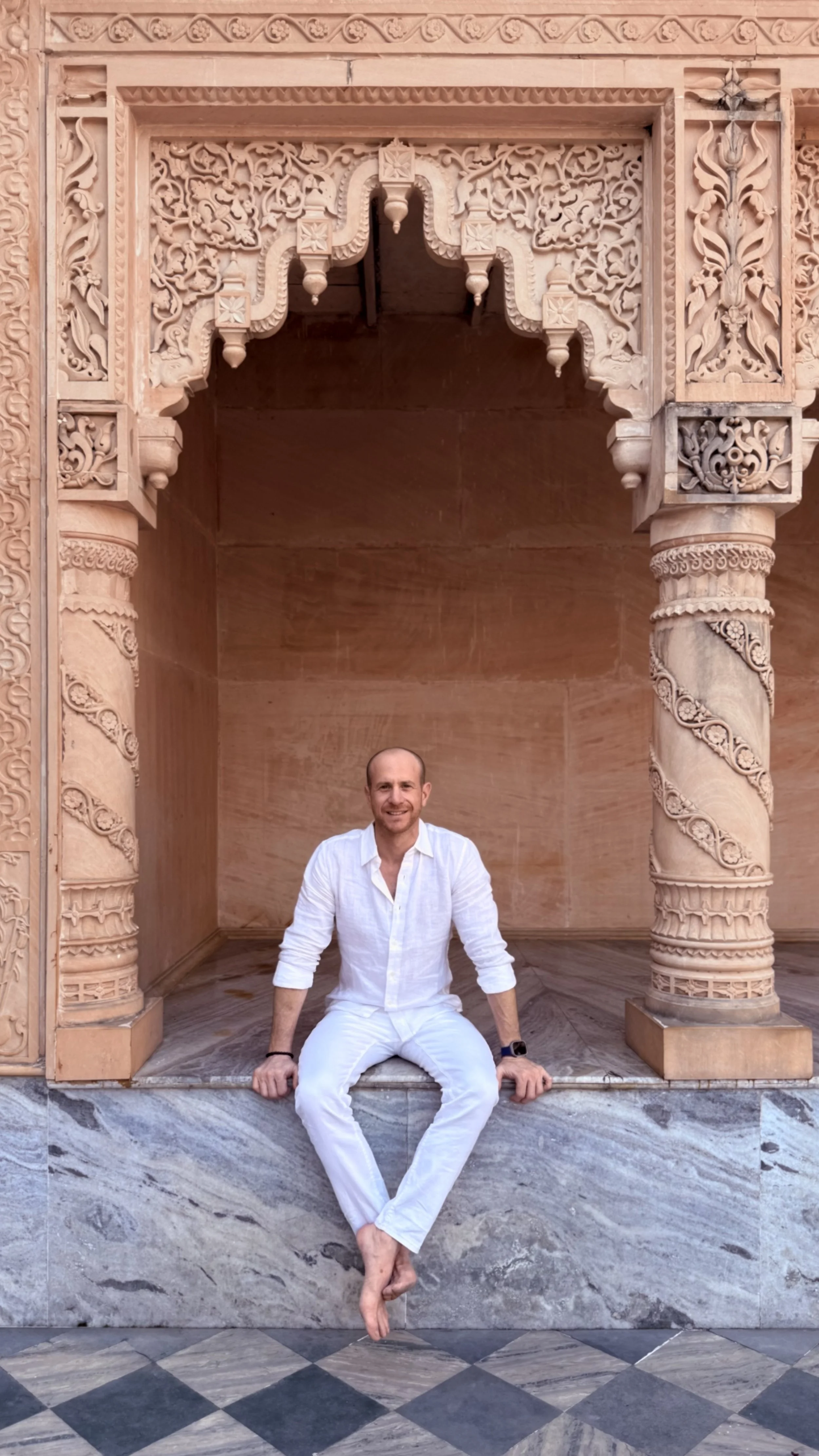 Person sitting under intricately carved stone archway, wearing white clothes, with patterned marble floor and ornate architectural details.
