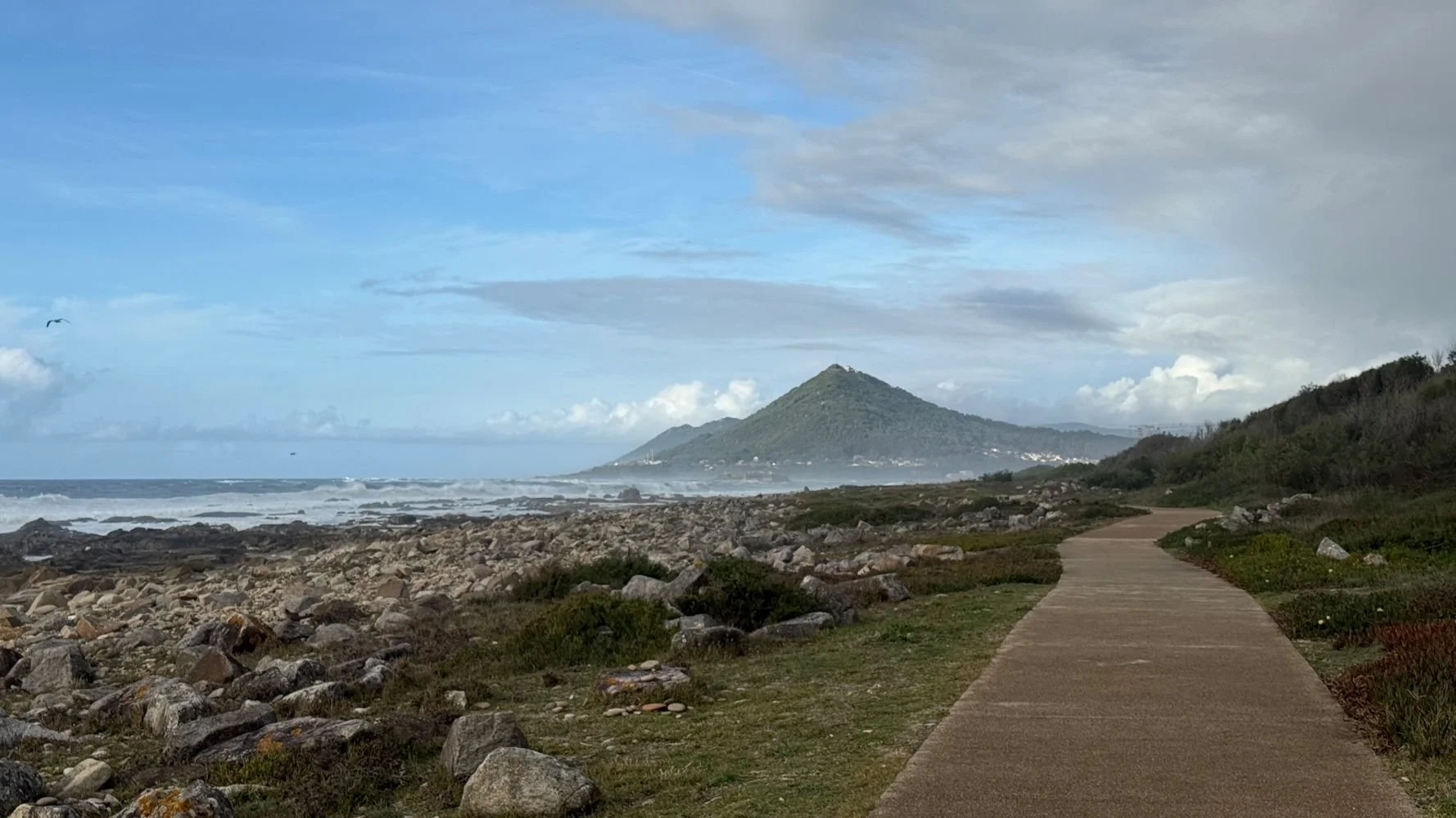 Coastal walking path on the Camino de Santiago in northern Spain, with the Atlantic Ocean on one side and a green mountain rising ahead