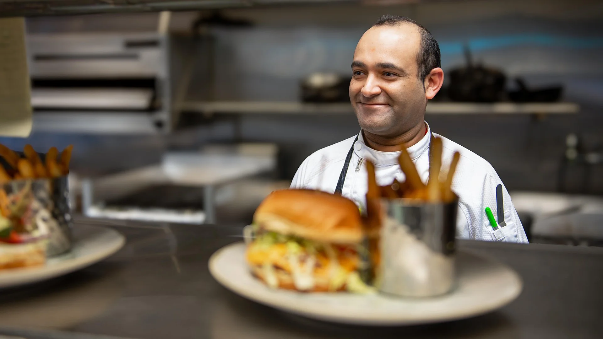 A chef in a kitchen looking at plates of burgers and fries.