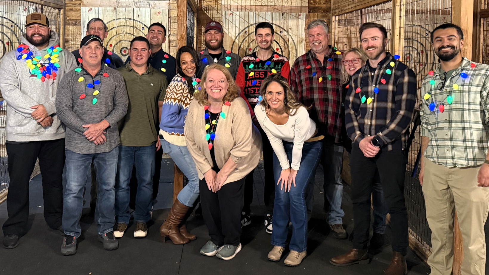 Group of people celebrating and smiling at an indoor activity, adorned with colorful Christmas lights and festive sweaters, with wooden wall background.