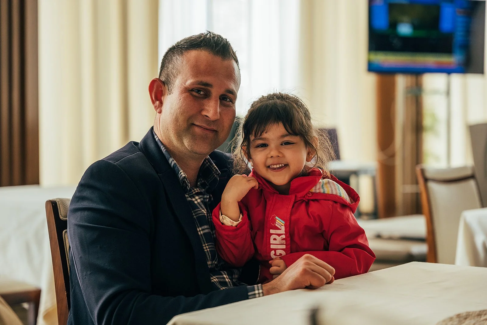 A man and young girl smiling while sitting at a table indoors, with a television screen and curtains in the background.