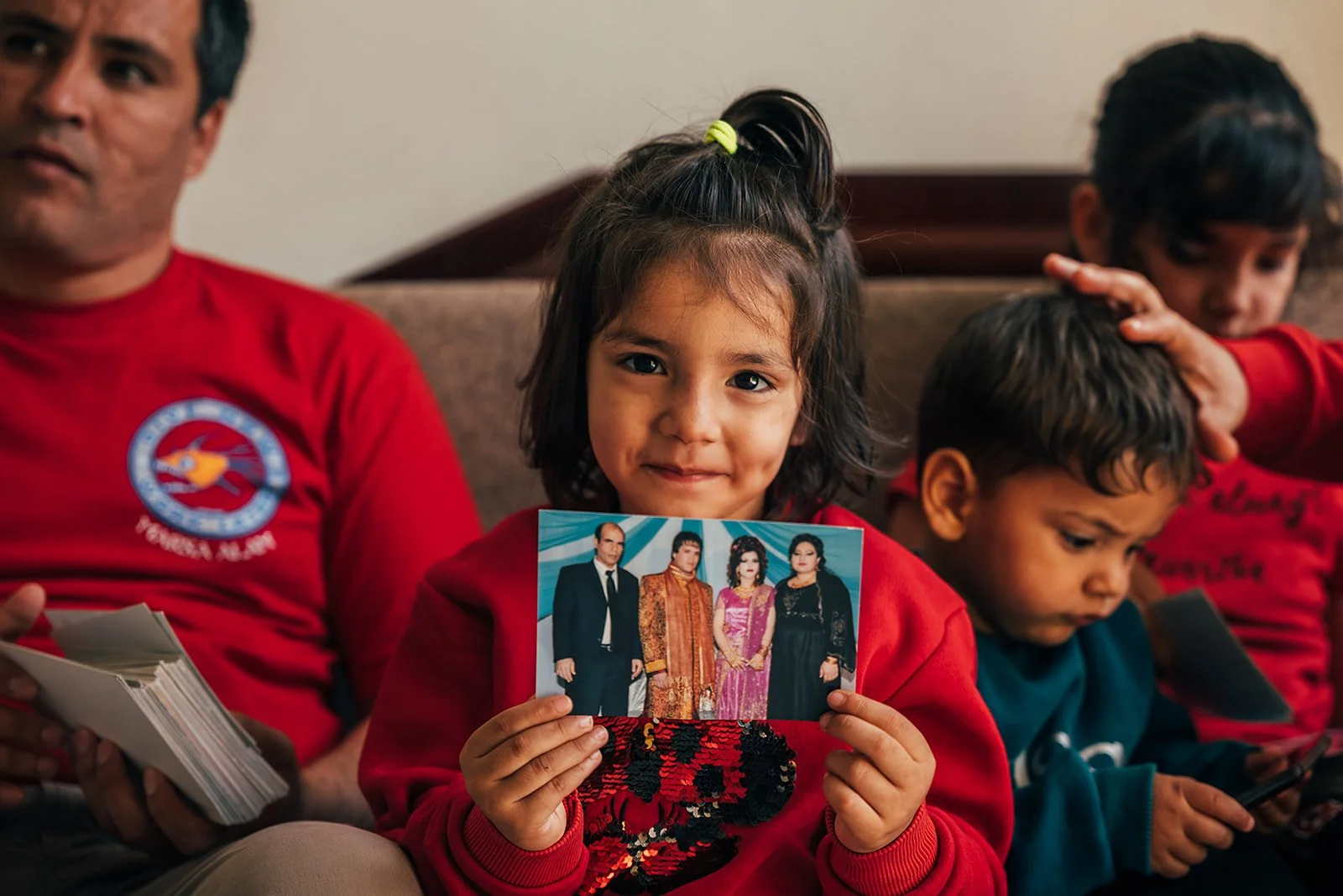 A young girl with dark hair in a ponytail, holding a Family photo, sitting on a couch with other children and an adult, all wearing red and teal shirts