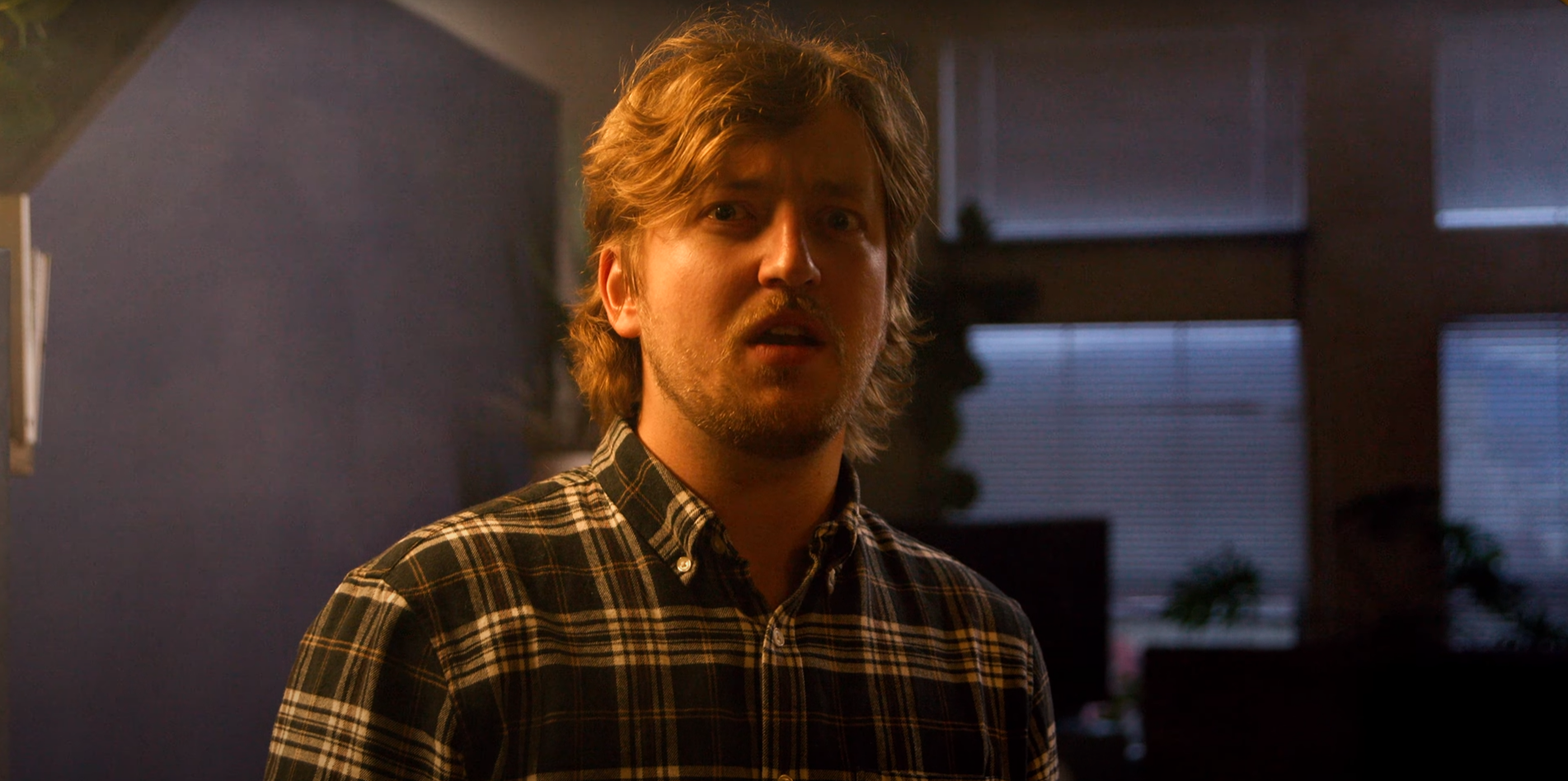 A man with medium-length, wavy blond hair and a beard, wearing a black, white, and yellow plaid shirt, standing indoors in a dimly lit room with blue blinds in the background.