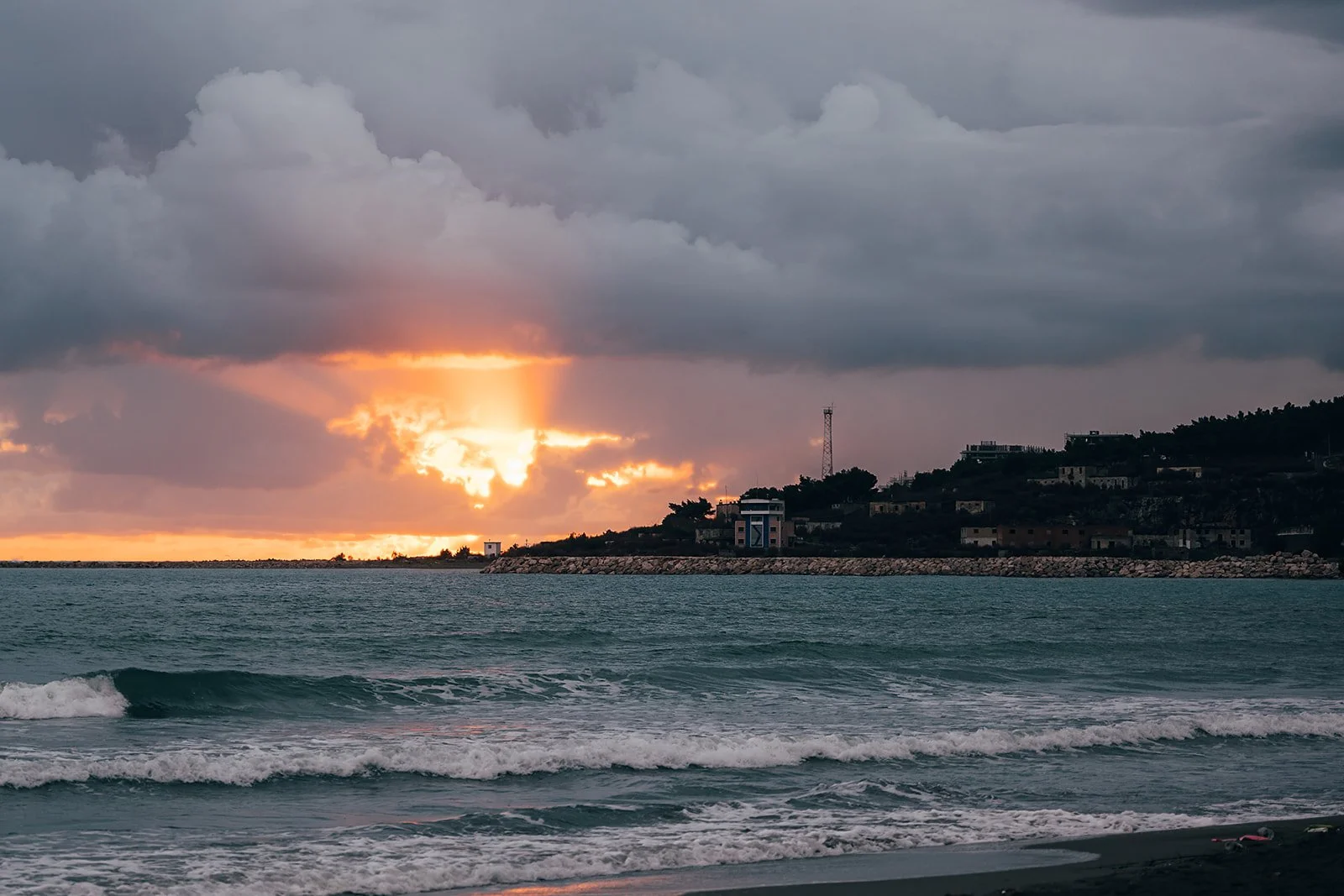 Sunset over the ocean with clouds and a distant shoreline featuring buildings and trees.