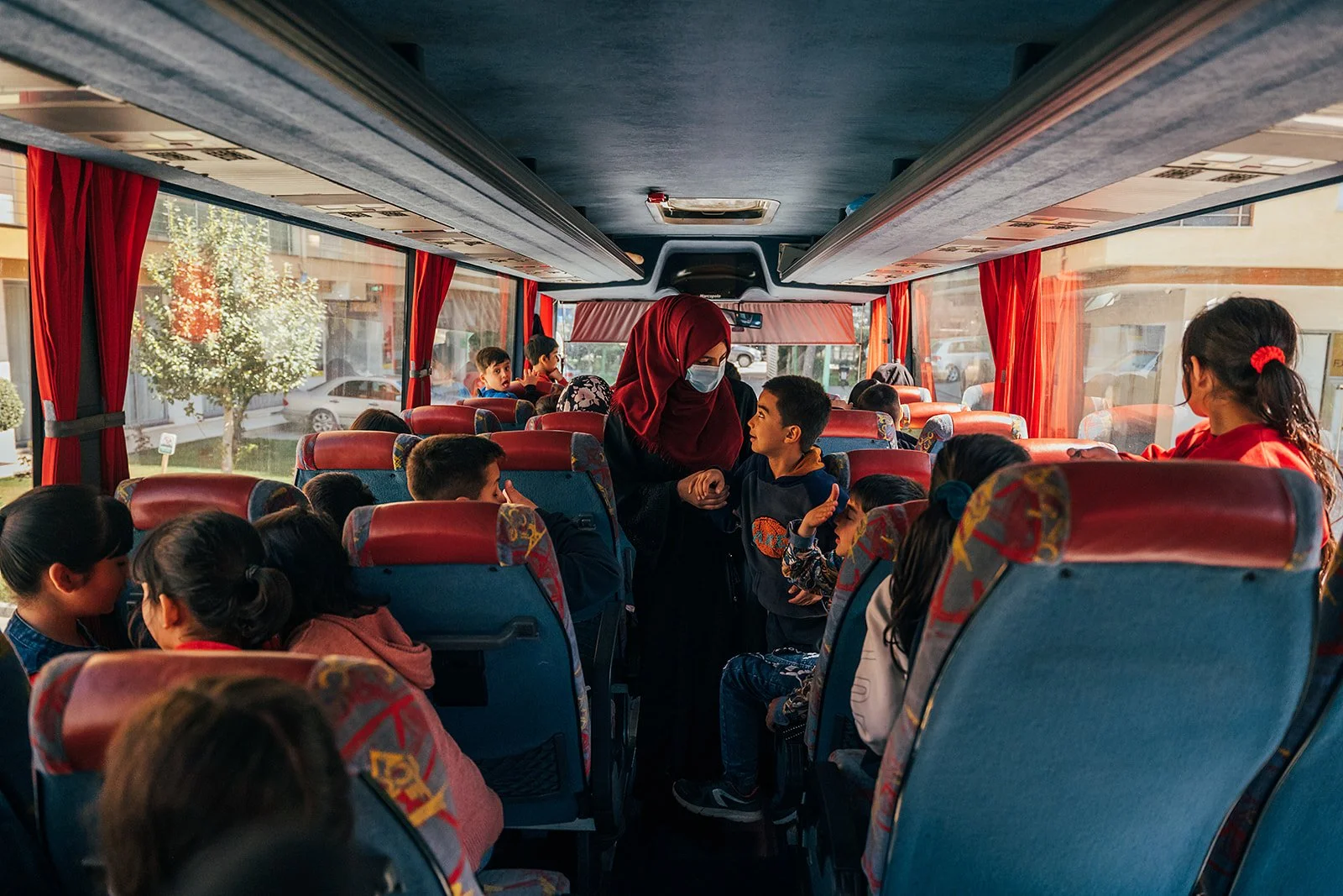 A group of children on a bus, with a woman in a red headscarf and face mask talking to a boy in the middle aisle.