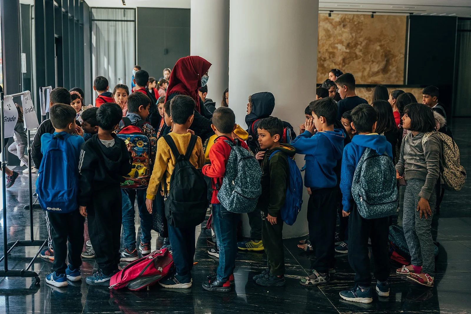 Group of school children with backpacks standing in line at an indoor location, possibly an airport or bus station, with a woman, likely a teacher or guardian, speaking to them.