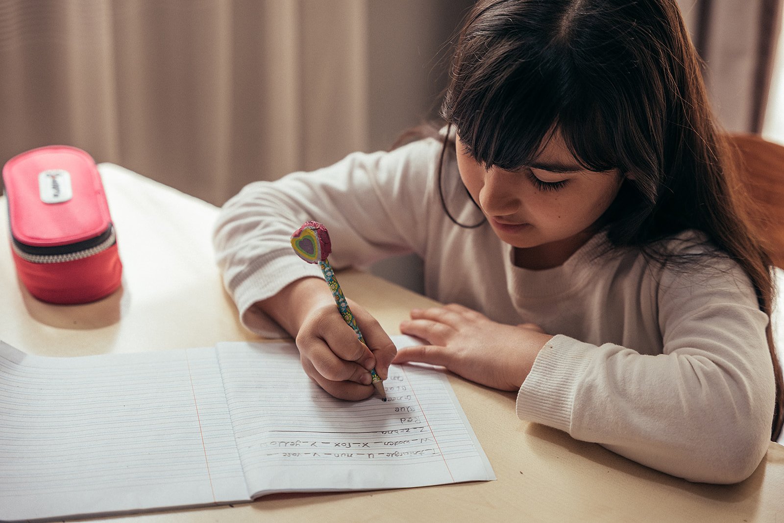 A young girl with dark hair and a white long-sleeve shirt sitting at a table, writing in a notebook with a colorful heart-patterned pencil, with a pink pencil case nearby.