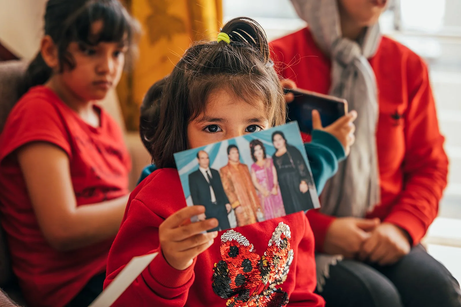 A young girl in a red jacket holding a photo of four people, with two other children and a woman sitting nearby, in a cozy indoor setting.