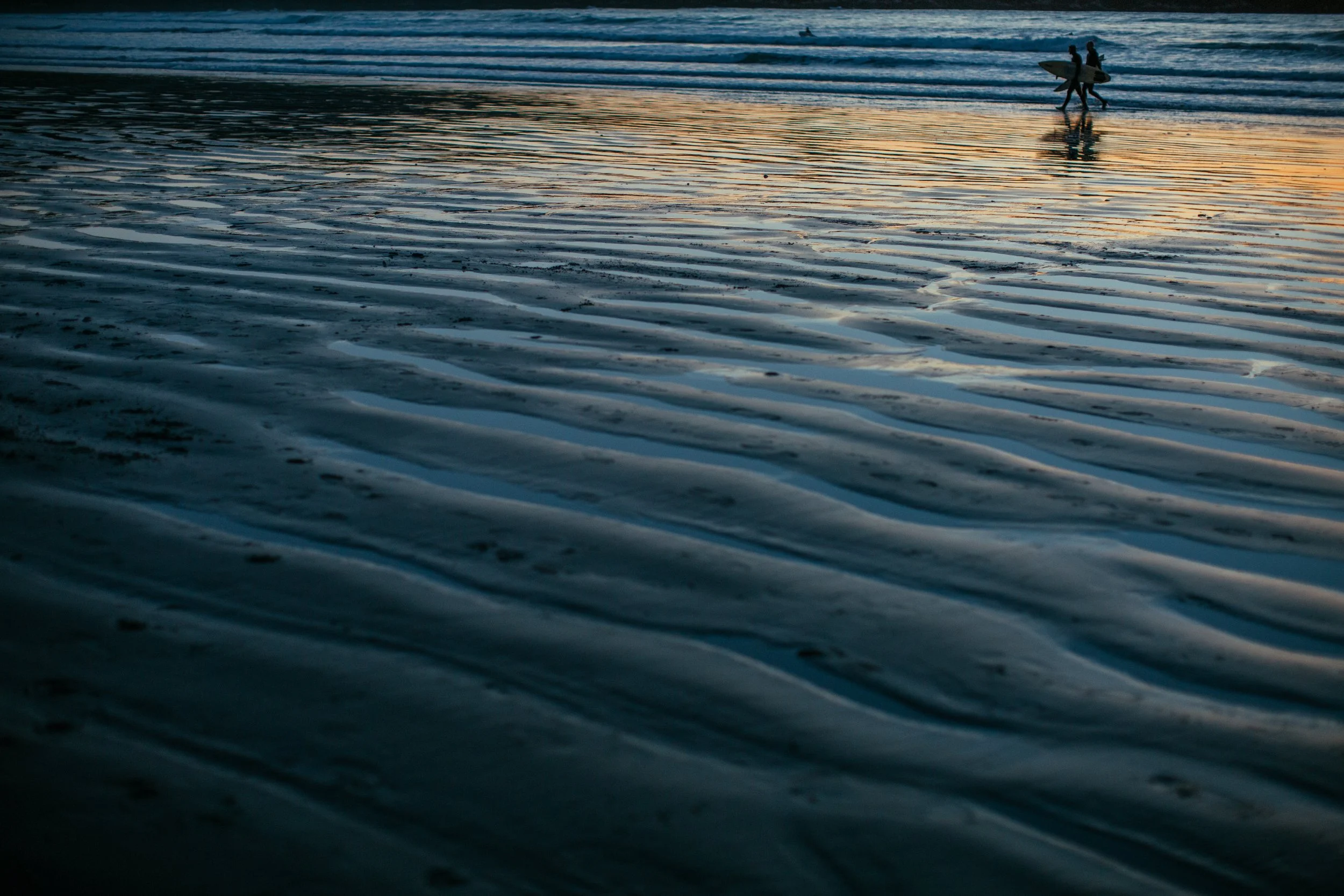 Two surfers walking on wet sand near the shoreline at sunset, carrying surfboards, with gentle waves in the background.