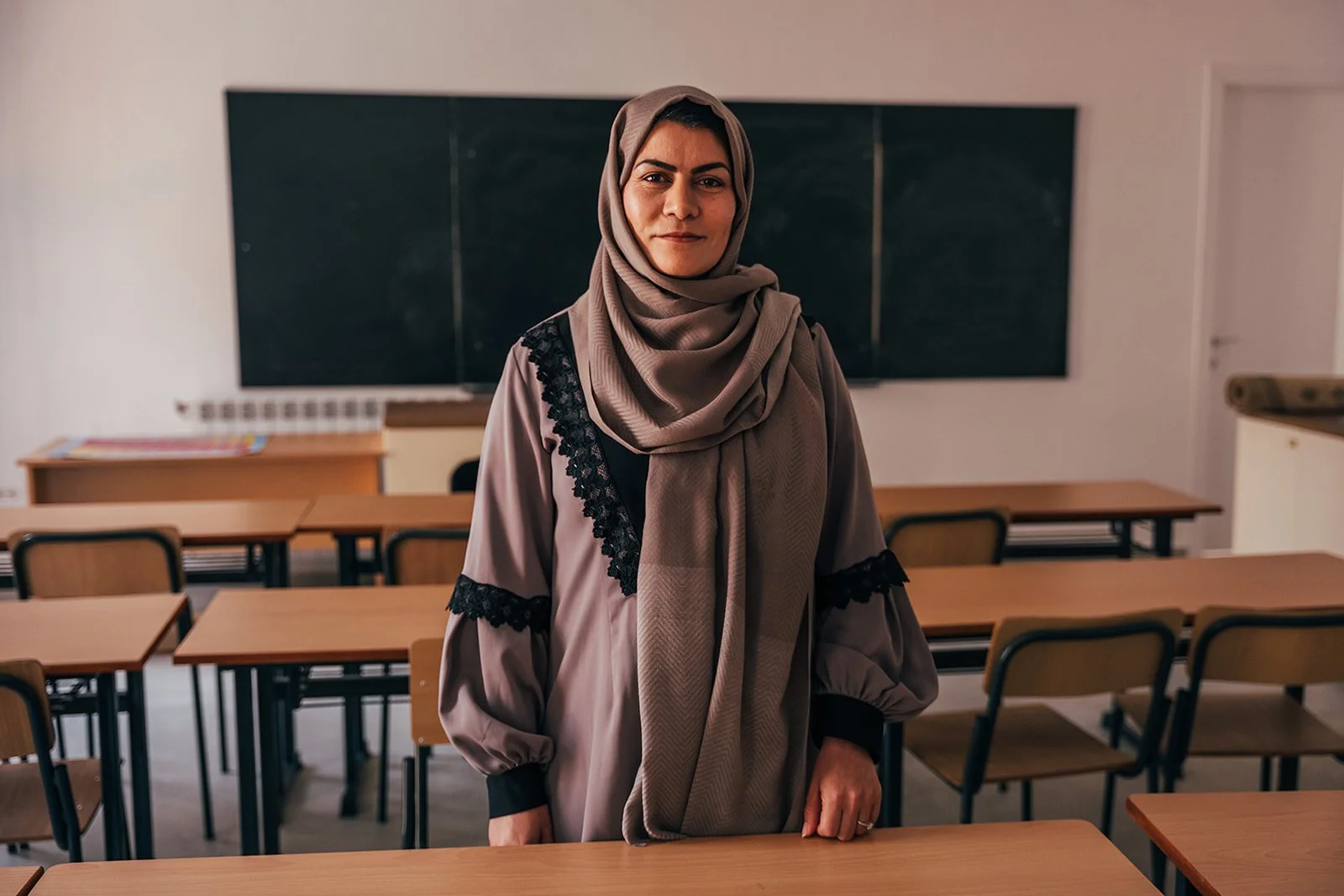 A woman wearing a hijab and a gray dress standing in a classroom with desks and chairs, a chalkboard in the background.