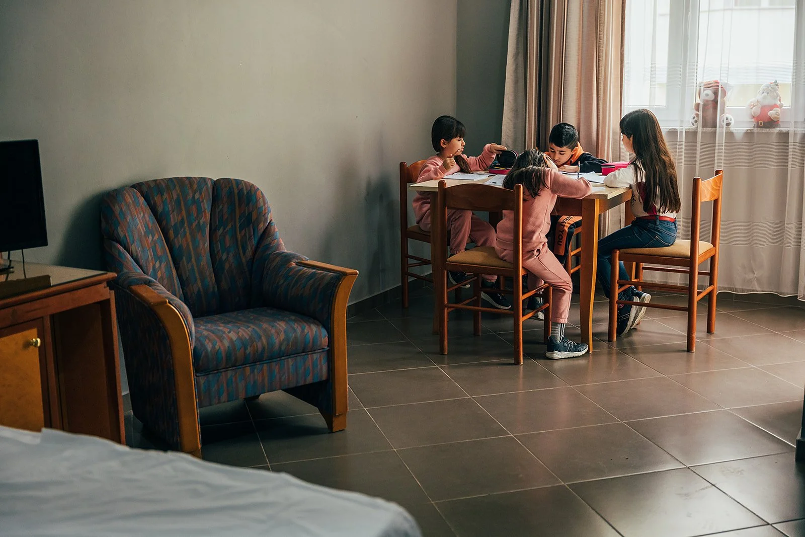 Four children sitting at a wooden dining table in a hotel room, working on homework or in a classroom setting, with a window and curtains in the background.