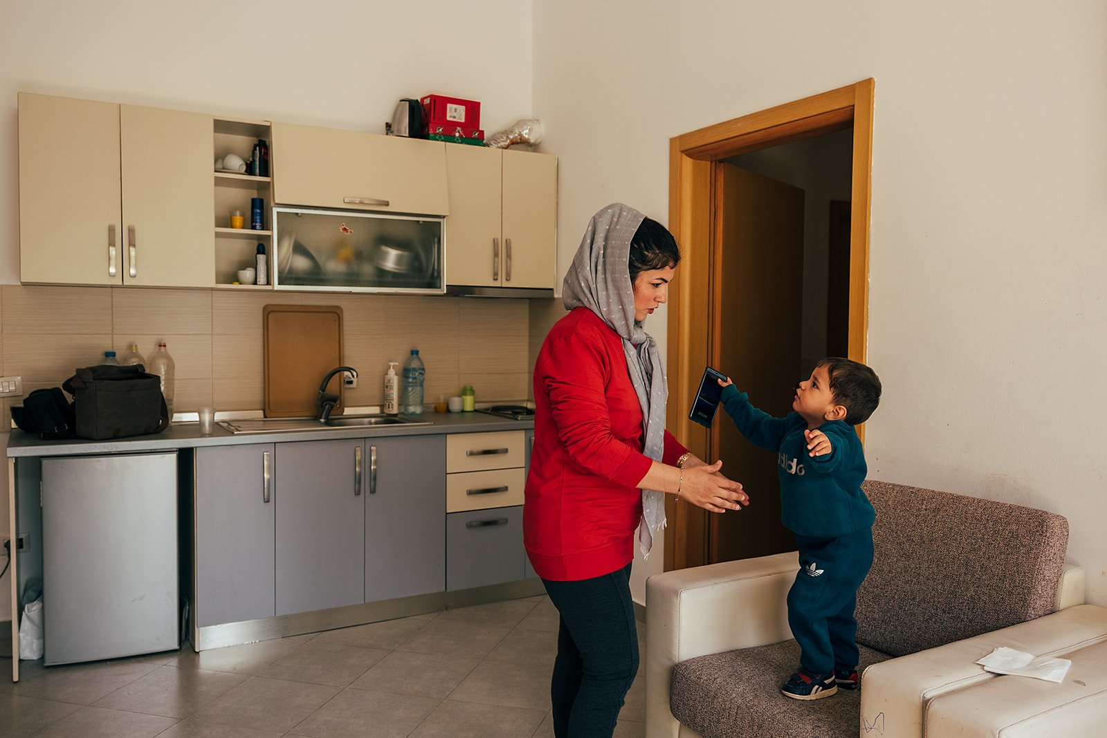 Woman in red shirt and gray headscarf interacting with young boy holding a phone in a kitchen with cream and gray cabinets and a brown door.