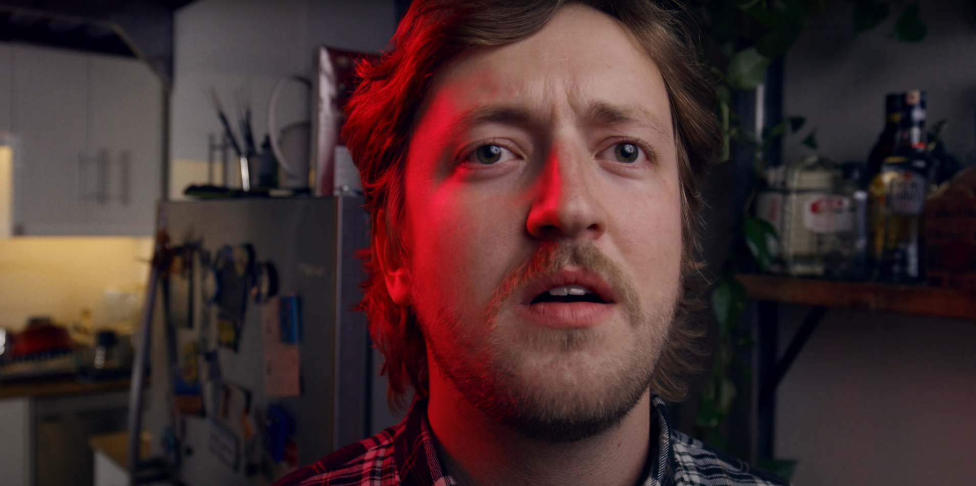 Close-up of a man with medium-length brown hair and a beard, looking surprised or confused in a kitchen with various appliances and bottles in the background.